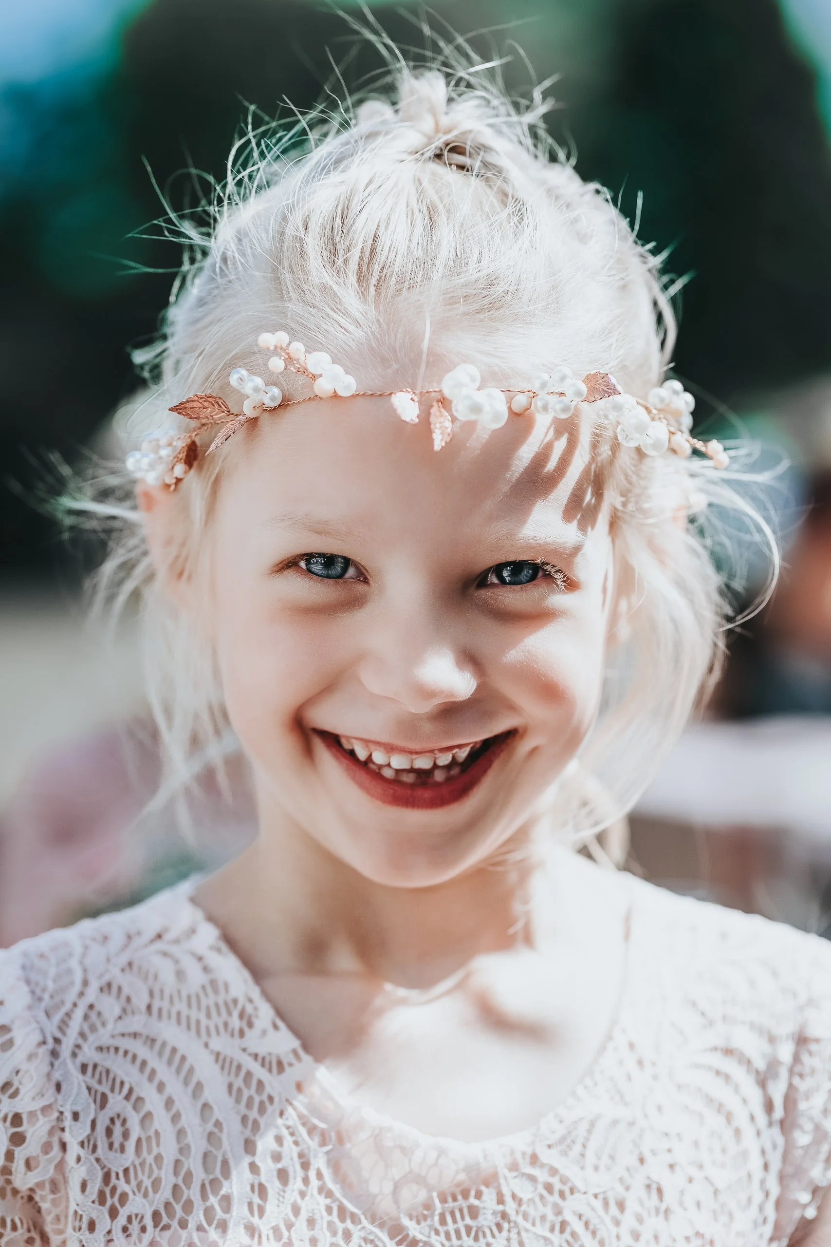 Ein lachendes Mädchen mit blonden, lockigen Haaren, trägt einen Blumenkranz und ein weißes Spitzenkleid.