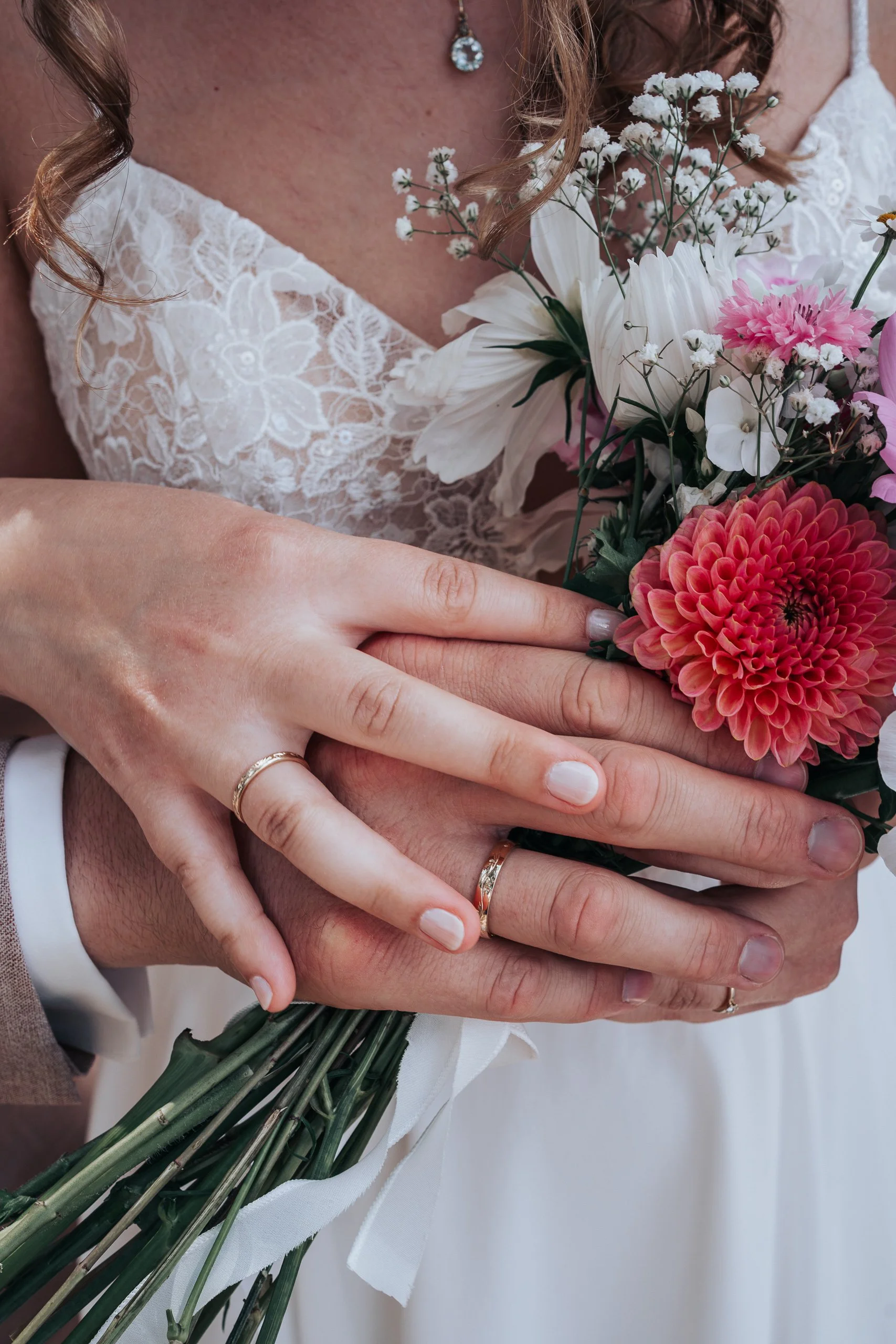 Nahaufnahme eines Brautpaares, das sich umarmt, mit einem Blumenstrauß aus weißen, rosa und roten Blumen, sowie goldene Eheringe an den Händen.
