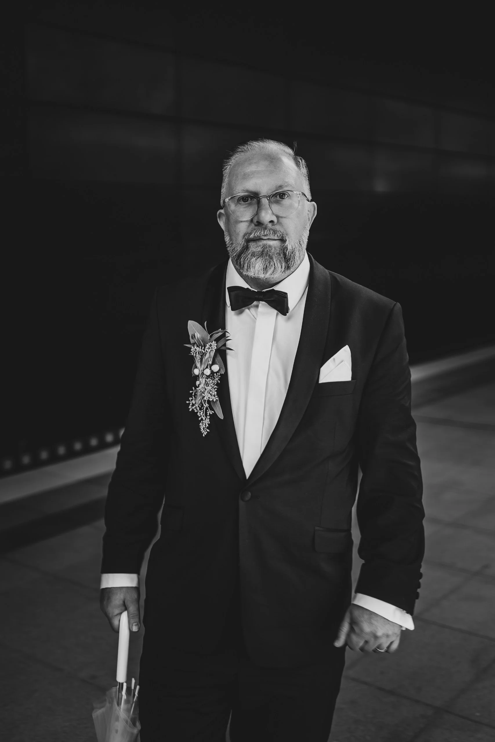 Schwarze und weiße Aufnahme eines Mannes in Smoking mit Fliege, Brille, Bart, der einen Blumenanstecker trägt und einen Regenschirm hält.