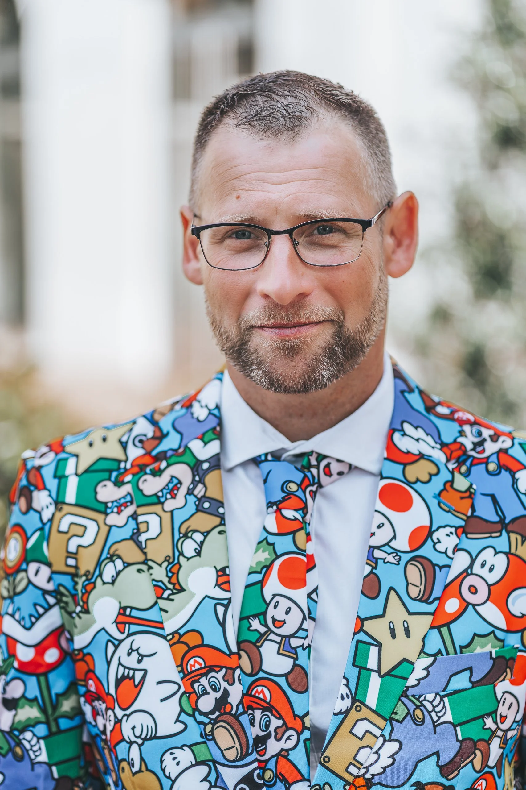 Ein Hochzeitsgast trägt einen bunten Anzug mit Super Mario Motiven, trägt eine Brille und hat einen kurzen, gepflegten Bart. Er steht draußen vor einem weißen Gebäude, blickt freundlich in die Kamera.