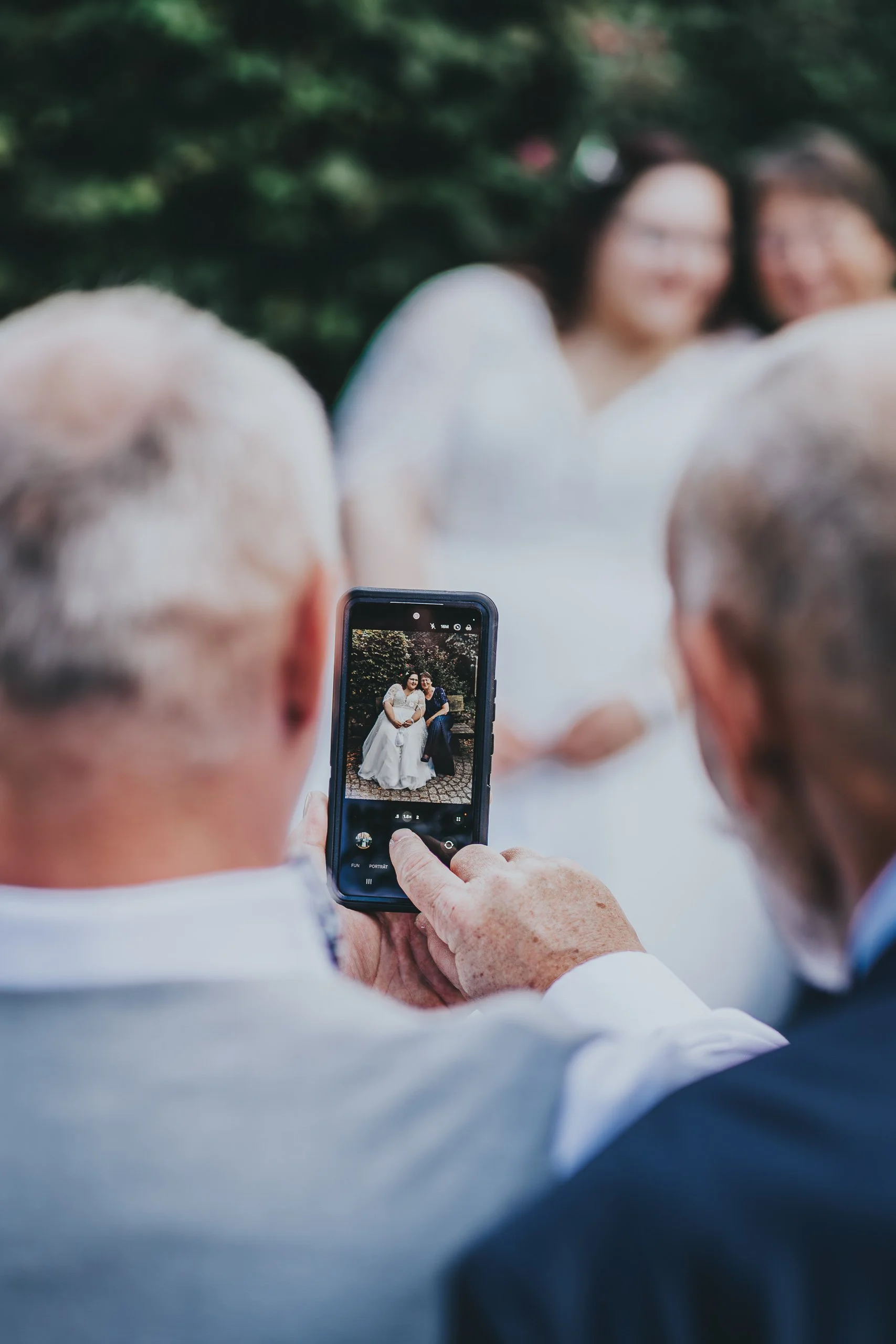 Zwei ältere Männer fotografieren ein Brautpaar auf einer Kamera in einem Park.