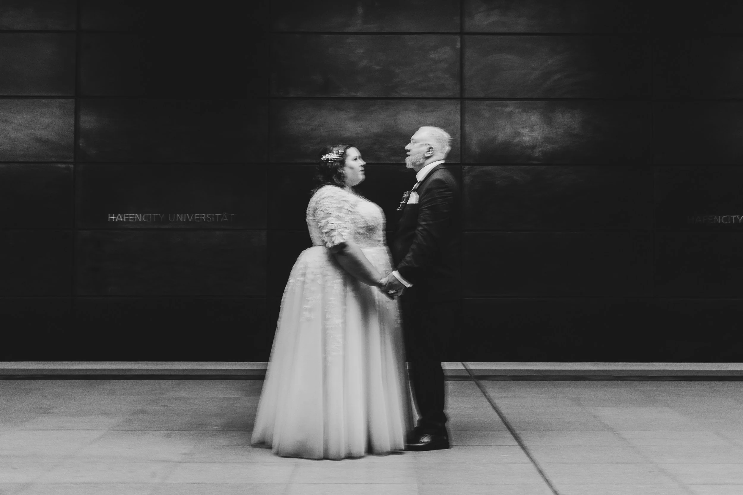 Verliebtes Paar in Hochzeitskleidung hält sich an den Händen vor schwarzer Wand mit der Aufschrift "HAFENCITY UNIVERSITÄT".