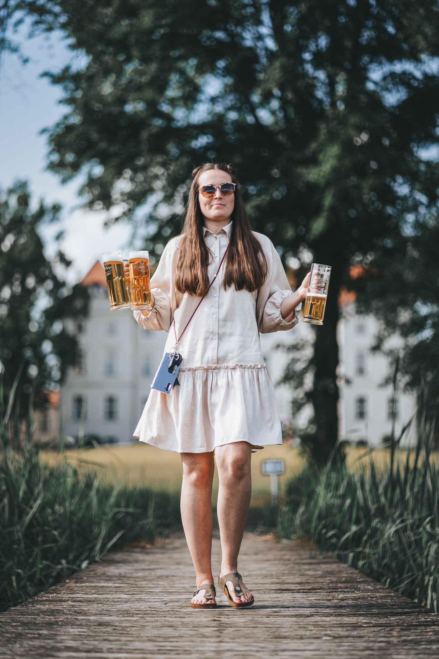 Eine junge Frau trägt Sonnenbrille, ein weißes Kleid, Sandalen und trägt mehrere Gläser Bier, während sie auf einem Holzsteg im Freien läuft.