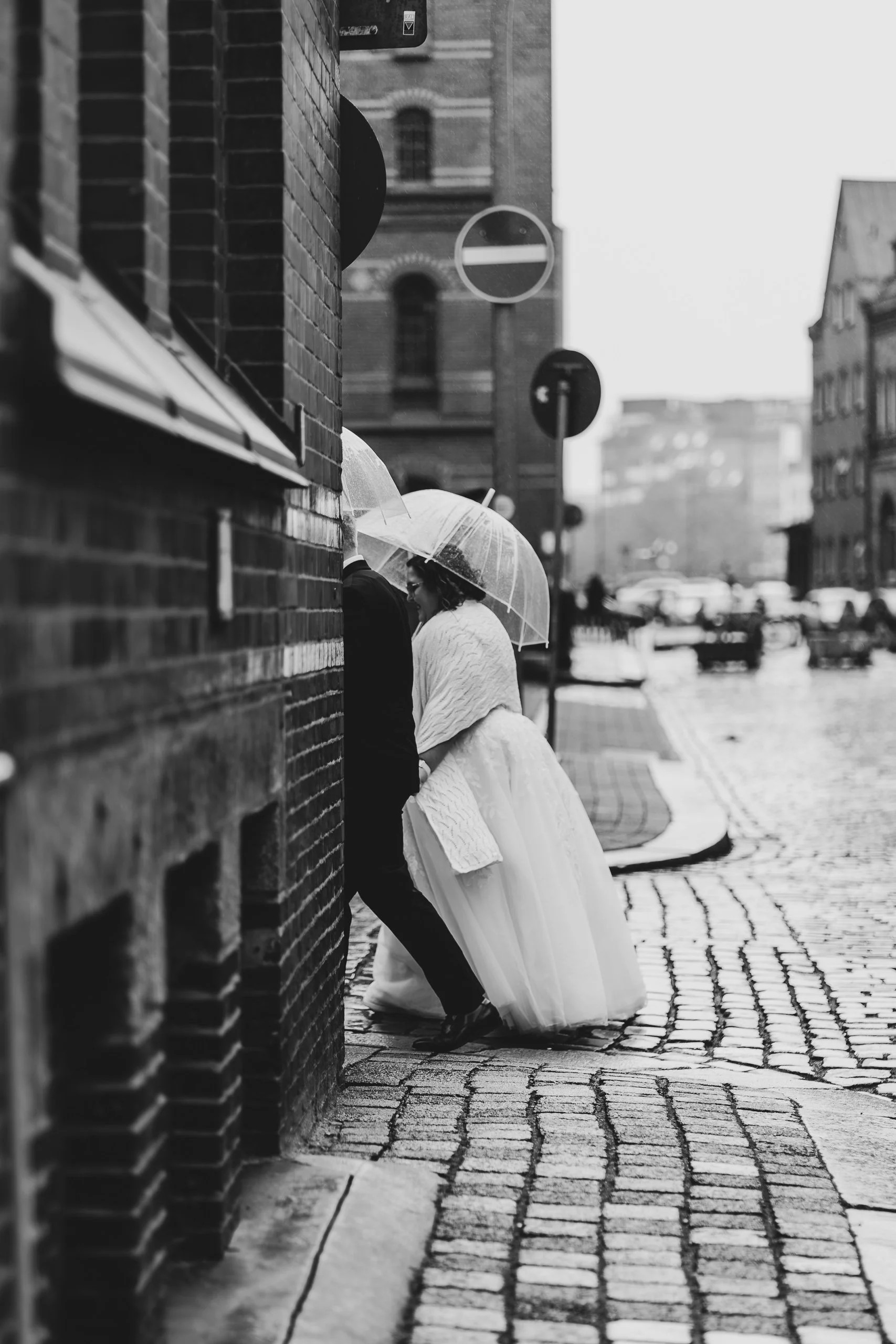 Paare bei einem Regenschauer auf einer gepflasterten Straße, mit Regenschirmen und einem Brautkleid.
