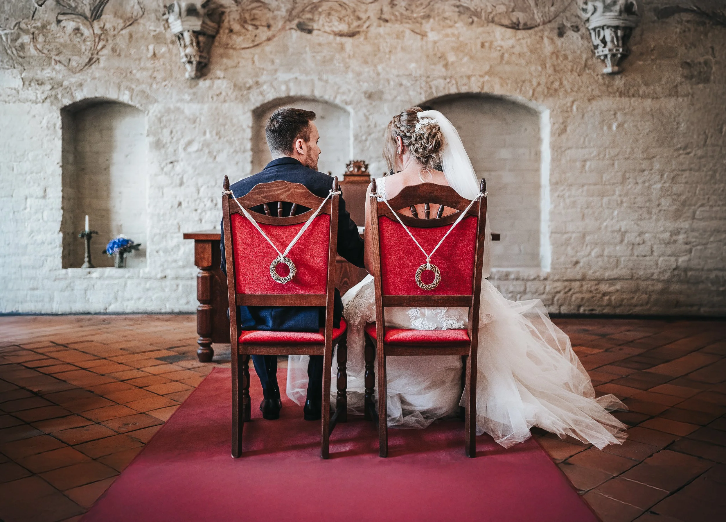 Braut und Bräutigam sitzen während ihrer standesamtlichen Trauung im Kloster Rehna nebeneinander auf roten, geschmückten Stühlen, mit Blick nach vorn vor weißen Ziegelwänden und historischem Interieur.