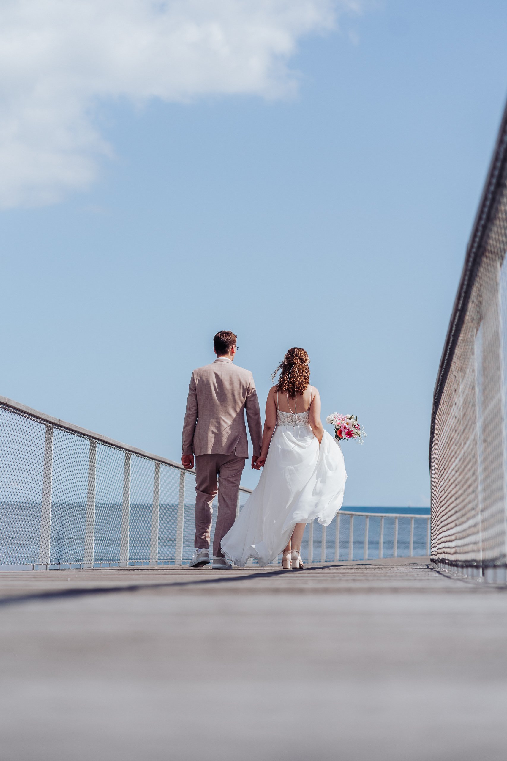 Brautpaar geht Hand in Hand über eine Brücke am Meer.