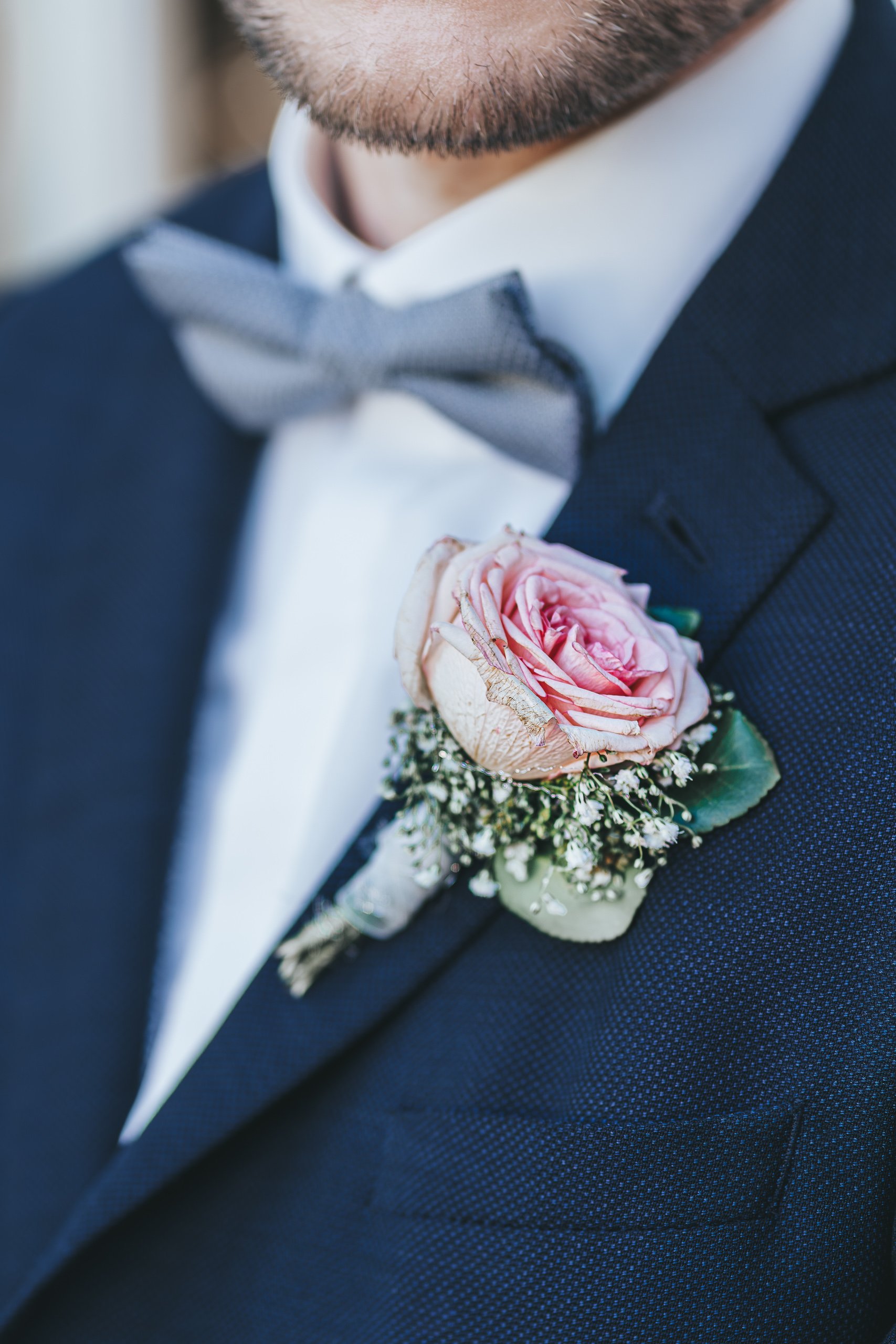 Nahaufnahme des Bräutigams in schwarzer Anzugjacke mit einer rosa-pinken Rose und weißen kleinen Blumen als Anstecker auf der linken Brustseite.