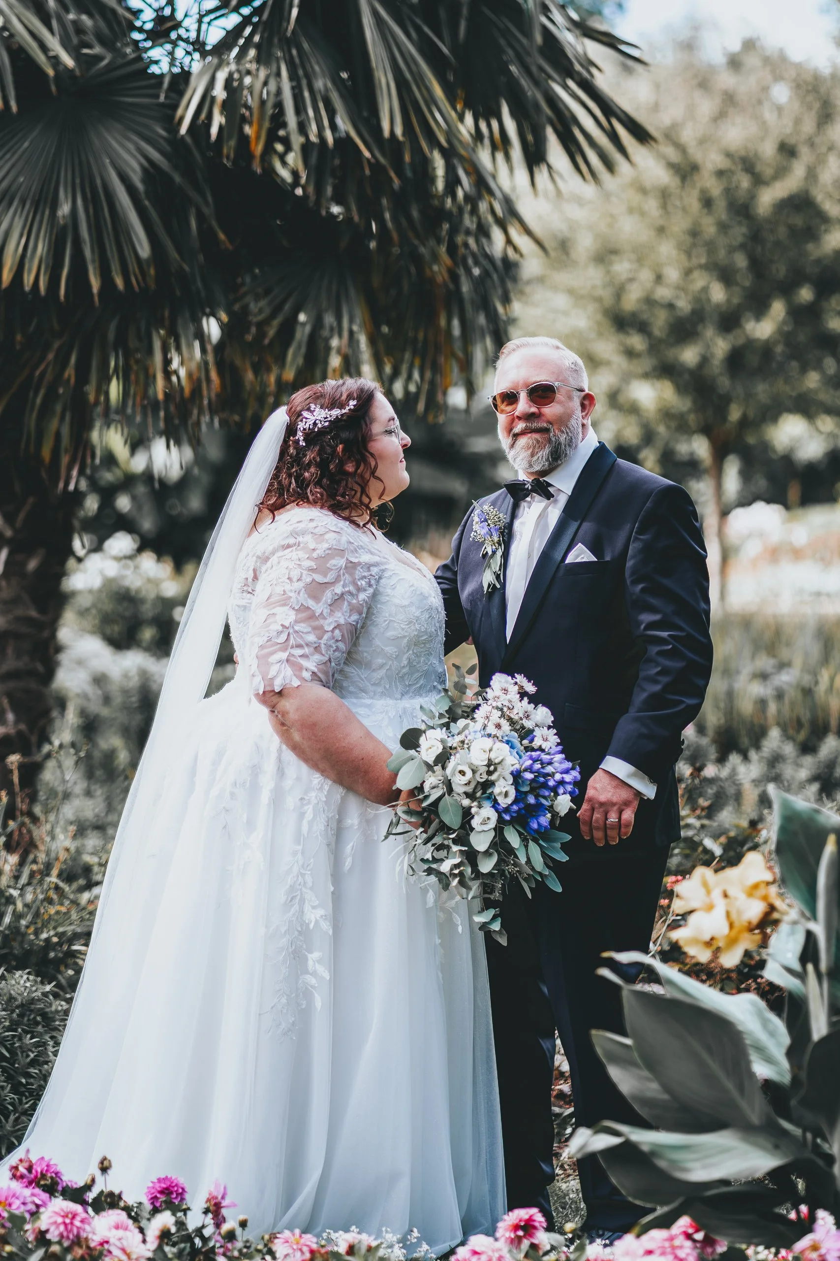 Ein Brautpaar bei ihrer Hochzeit im Freien, umgeben von Pflanzen und Blumen, die Braut trägt ein weißes Kleid mit Spitzen und einen Schleier, der Bräutigam trägt einen dunklen Anzug mit Fliege und Sonnenbrille, beide schauen sich liebevoll an.