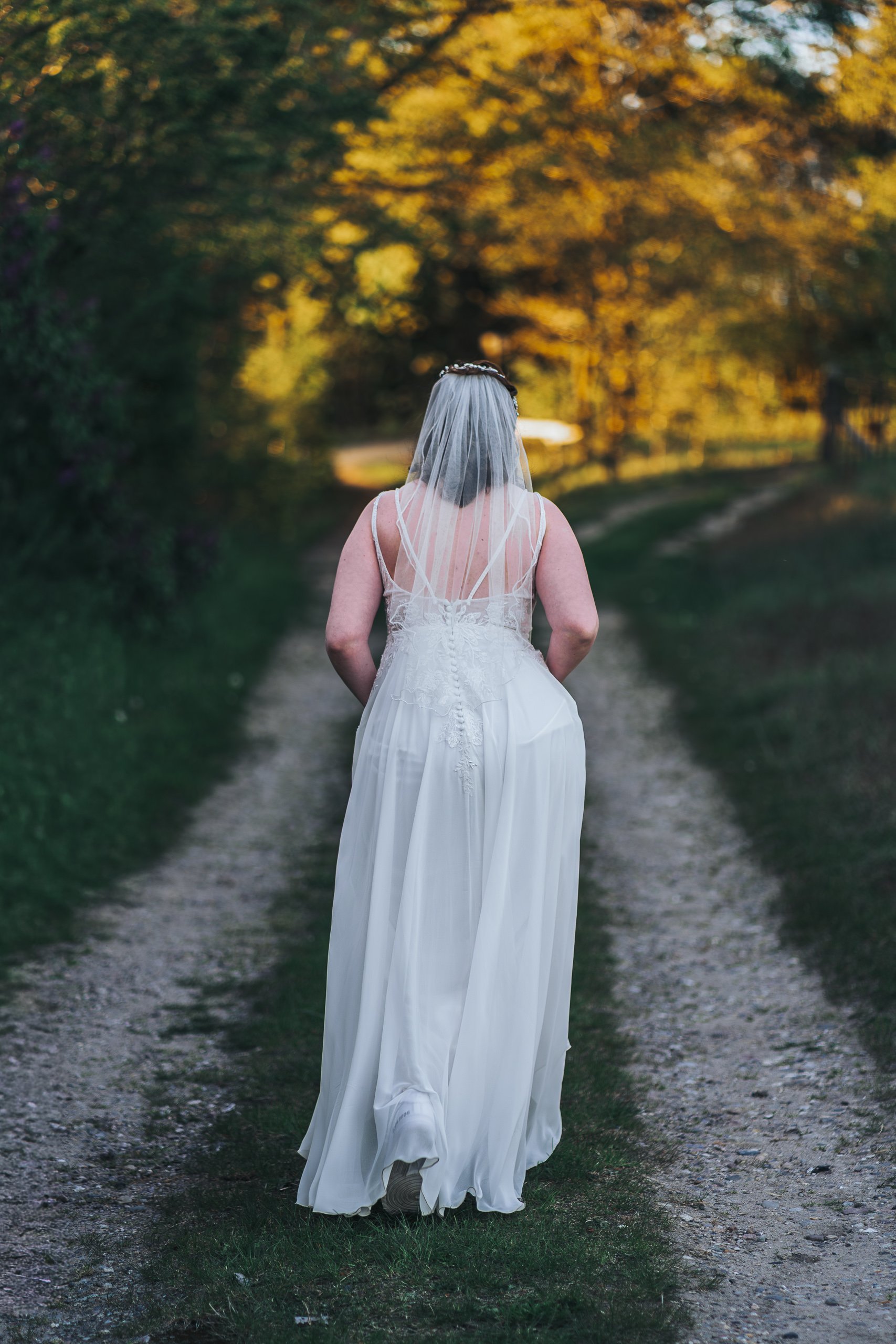 Eine Frau in einem weißen Hochzeitskleid mit Schleier läuft auf einem Wanderweg in einem Wald bei Sonnenuntergang.