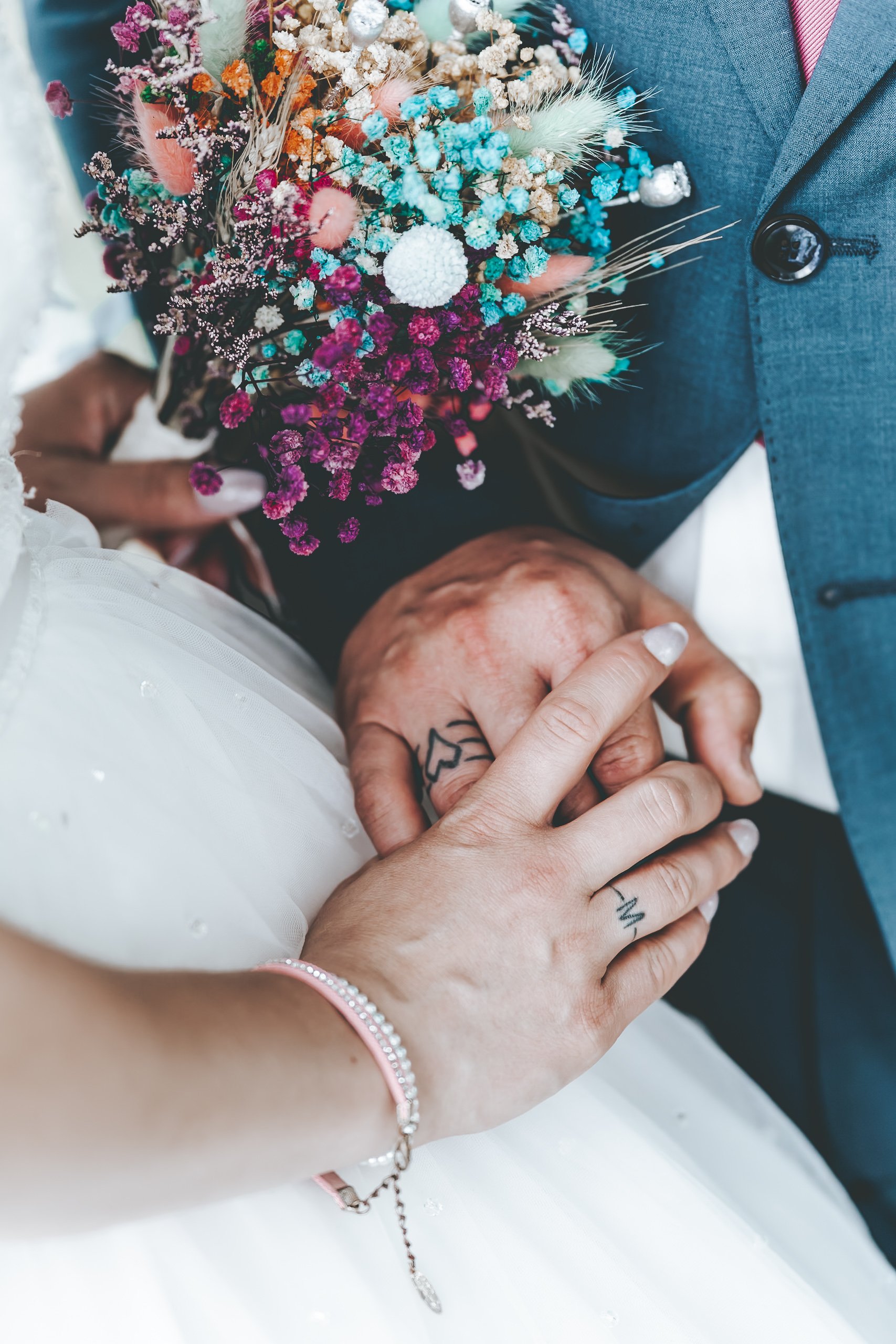 Ein Brautpaar hält Händchen, die Braut trägt ein weißes Kleid mit Perlen, eine Hand hat ein Tattoo mit einem Herz, das andere eine Armband. Die Braut hält einen bunten Blumenstrauß aus Trockenblumen, während der Bräutigam einen blauen Anzug trägt.