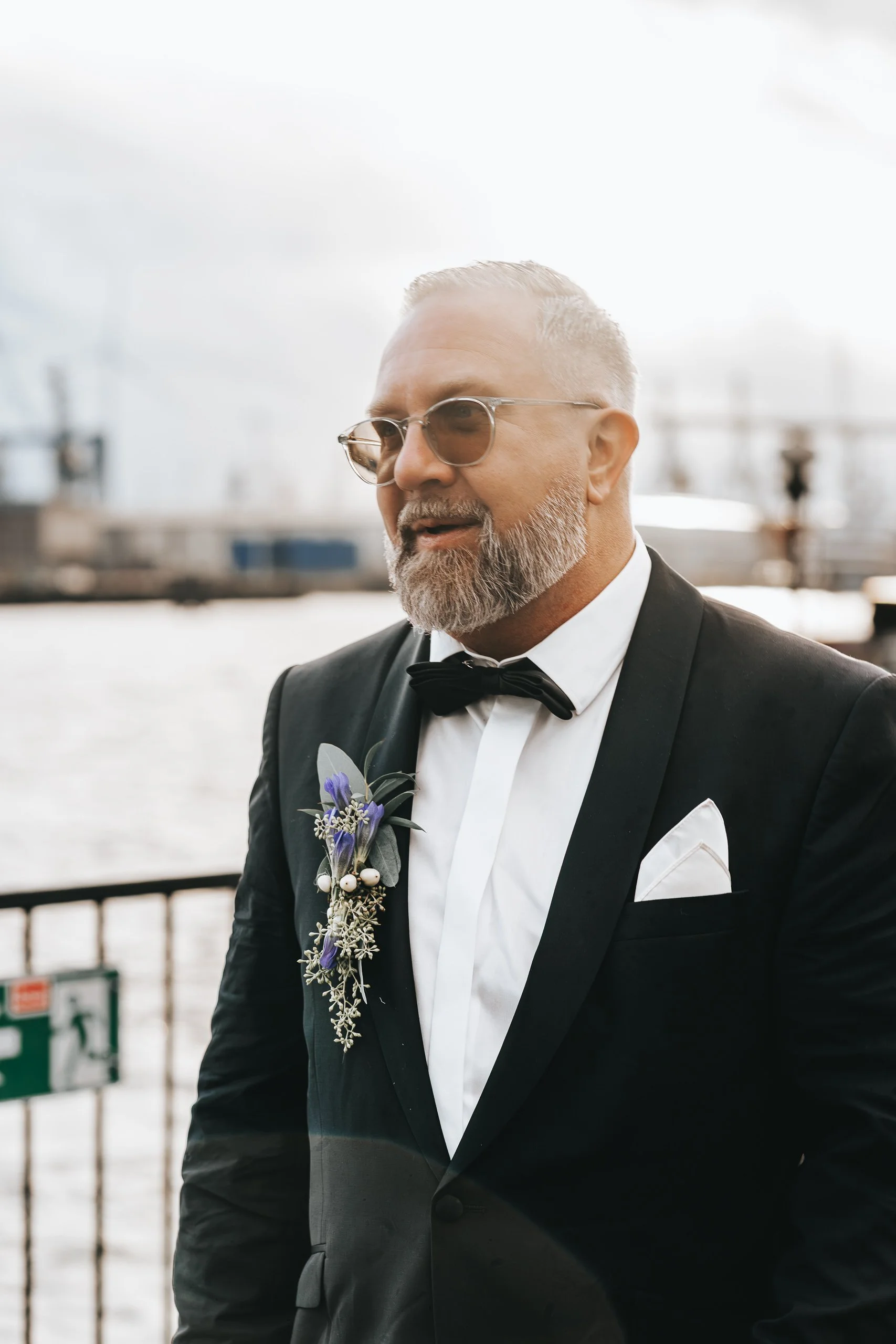Ein Mann in einem schwarzen Smoking mit Fliege und weißem Hemd, trägt eine Blumenbrosche am Revers, steht neben einem Wasserweg mit Hafen und Brücke im Hintergrund, trägt eine Sonnenbrille.