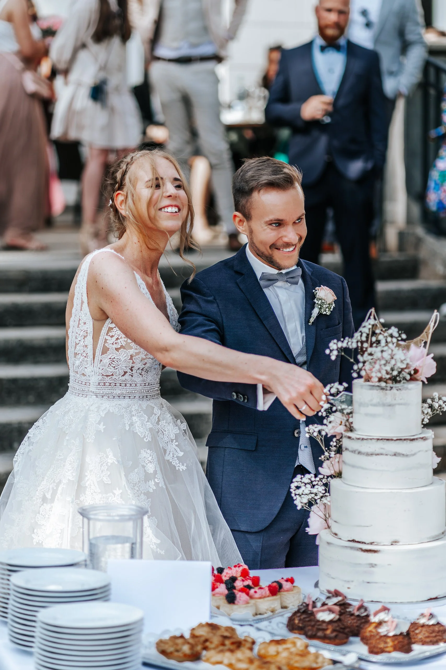 Braut und Bräutigam schneiden eine Hochzeitstorte bei einer Feier im Freien, im Hintergrund feiern Gäste.