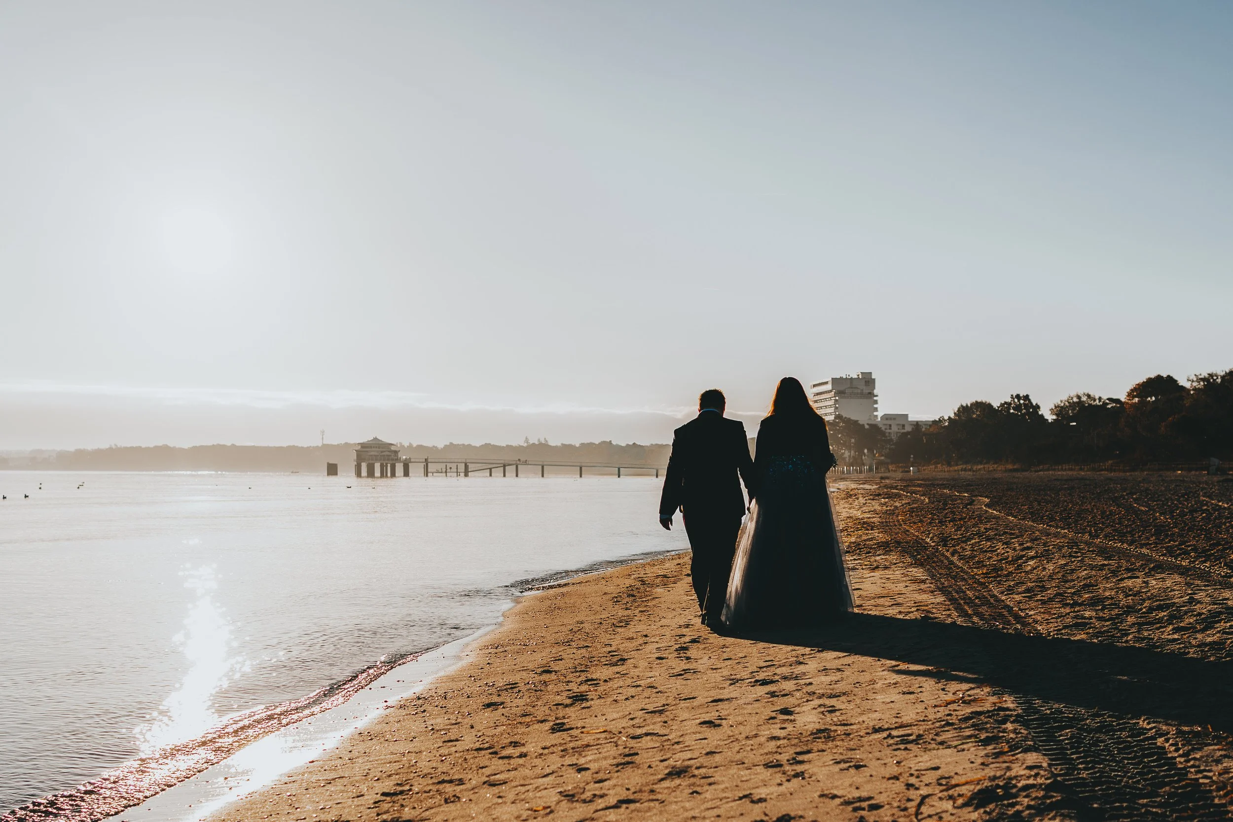 Ein Paar in formeller Kleidung spaziert am Strand bei Sonnenuntergang, der Himmel ist hell und die Szene ist ruhig.