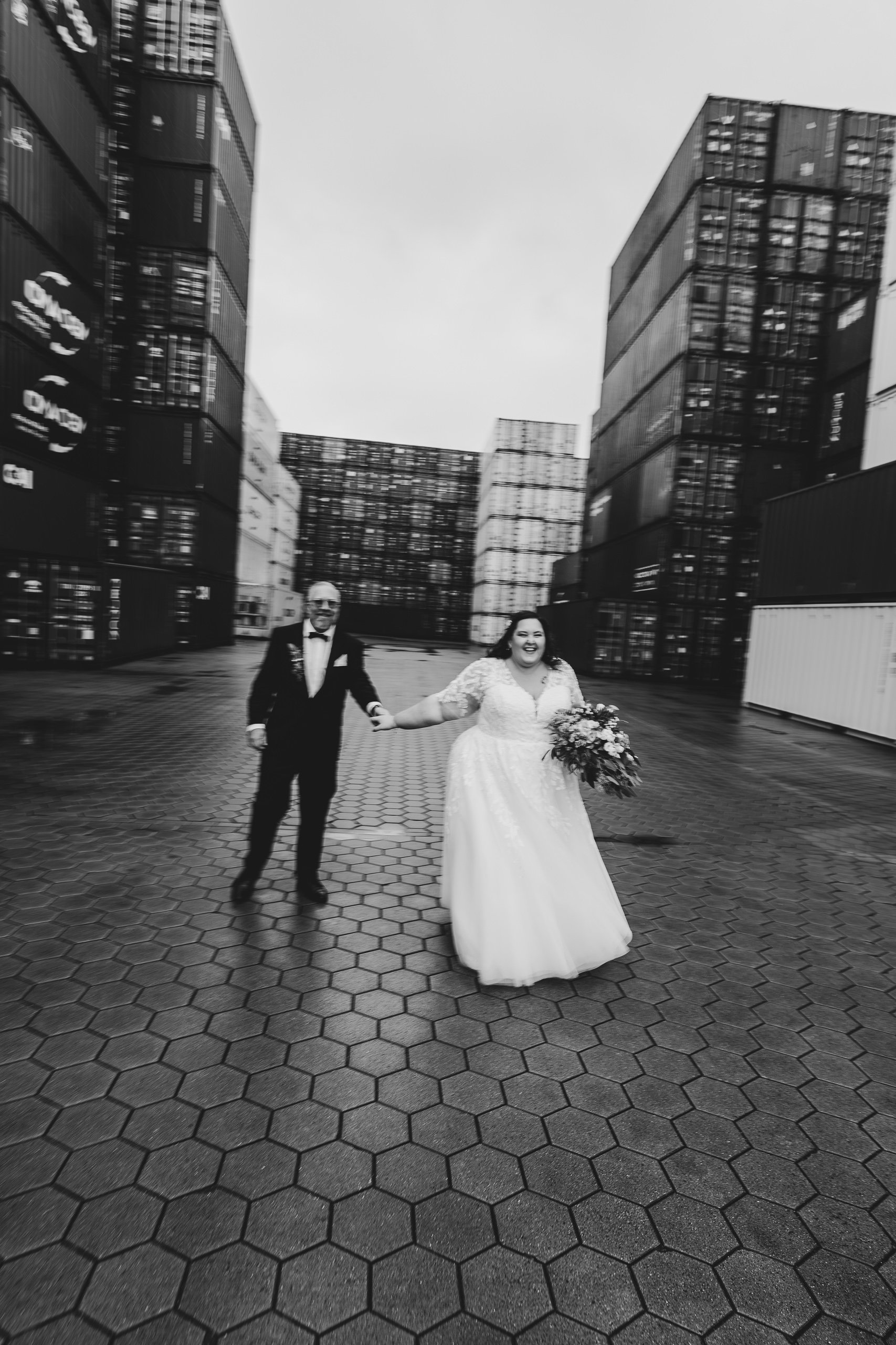 Ein Brautpaar in Hochzeitskleidung hält sich an einer industriellen Lagerhalle mit Containerregalen die eine urbane Atmosphäre schaffen, schwarz-weiß Aufnahme.