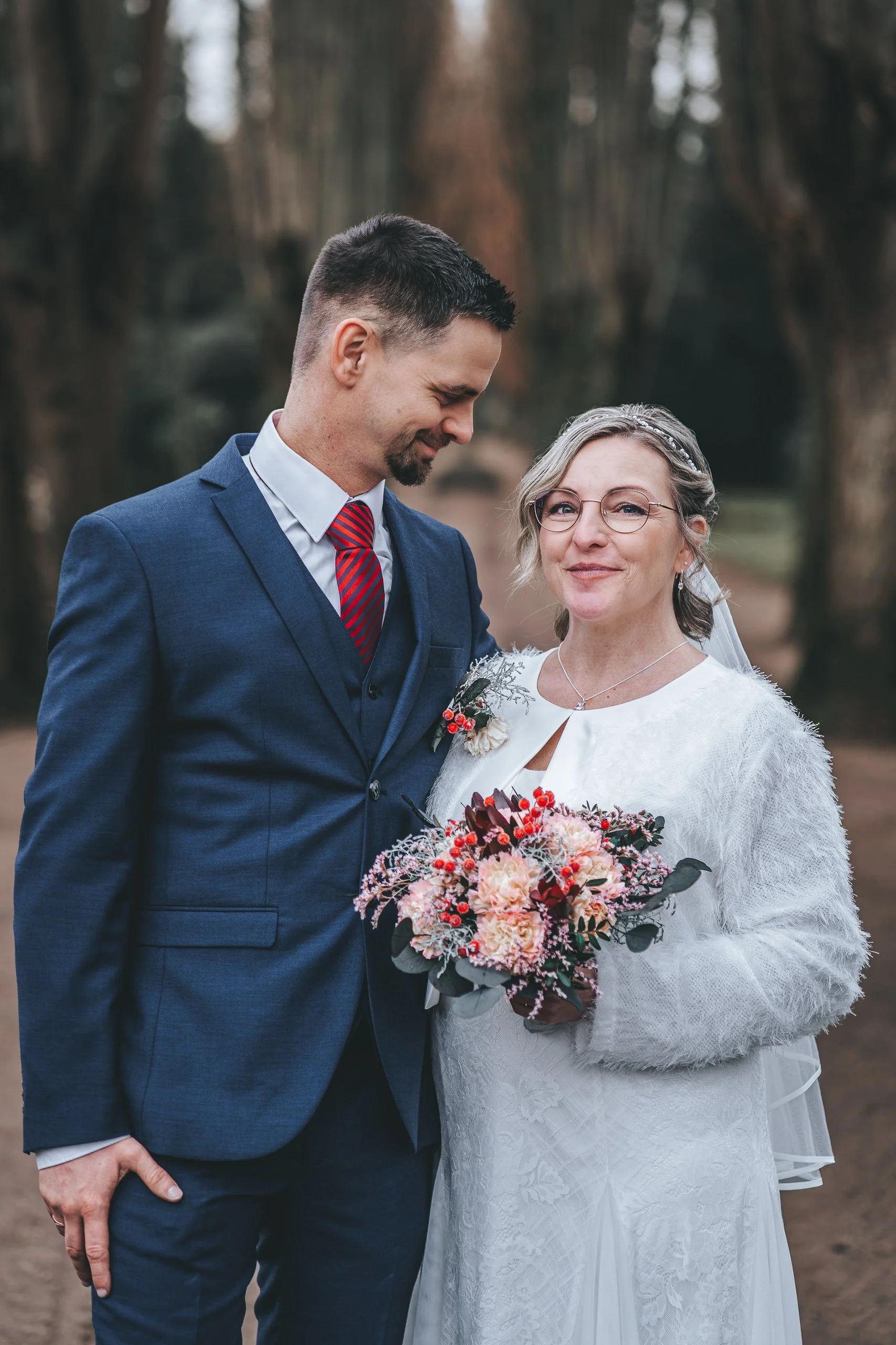 Brautpaar bei einer Winterhochzeit in einer Baumallee, Braut mit Strauß.