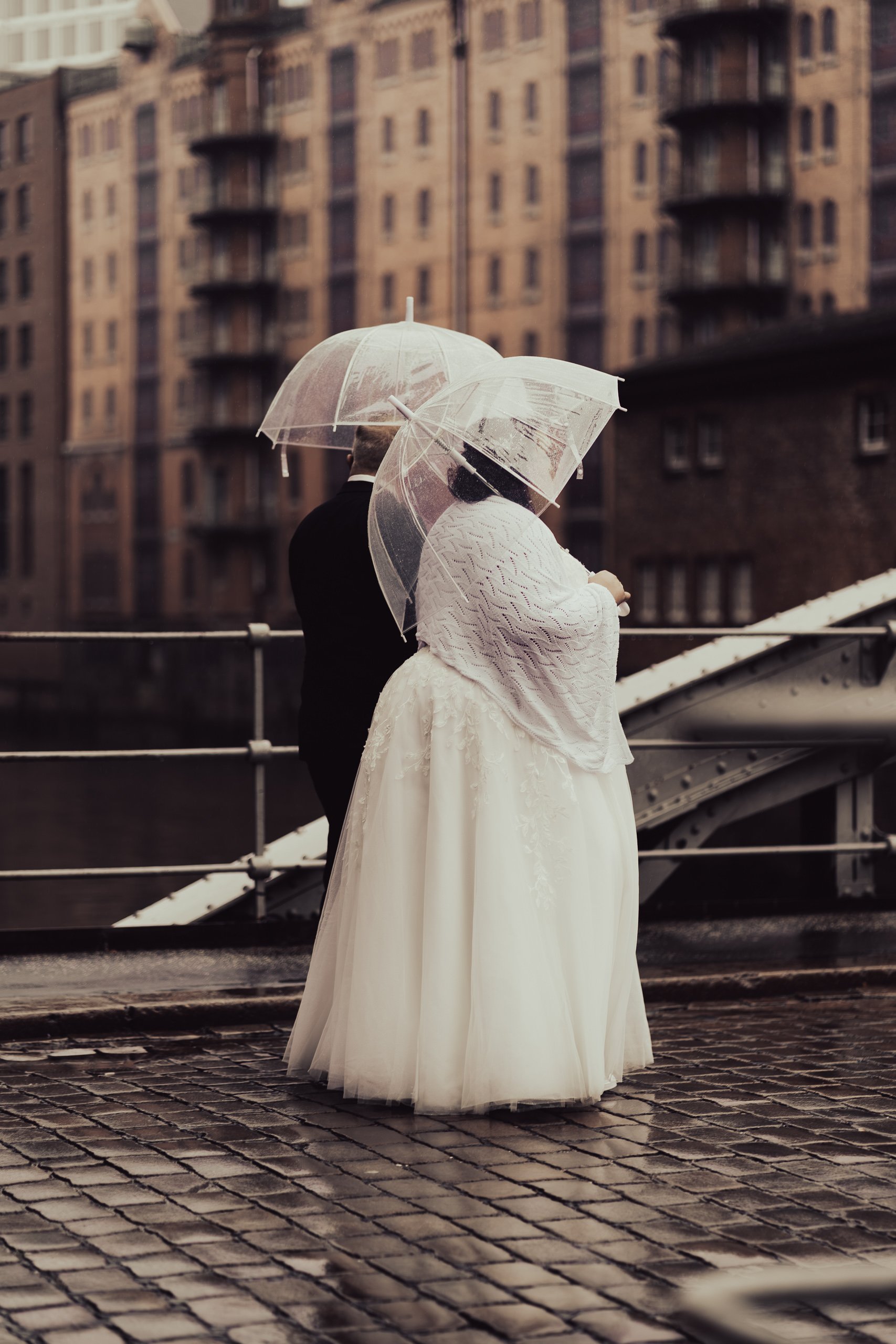 Ein Brautpaar mit Regenschirmen steht auf einem Kopfsteinpflaster vor einem Fluss, im Hintergrund sind Hochhäuser und eine Brücke, bei Regen, an einem grauen Tag.