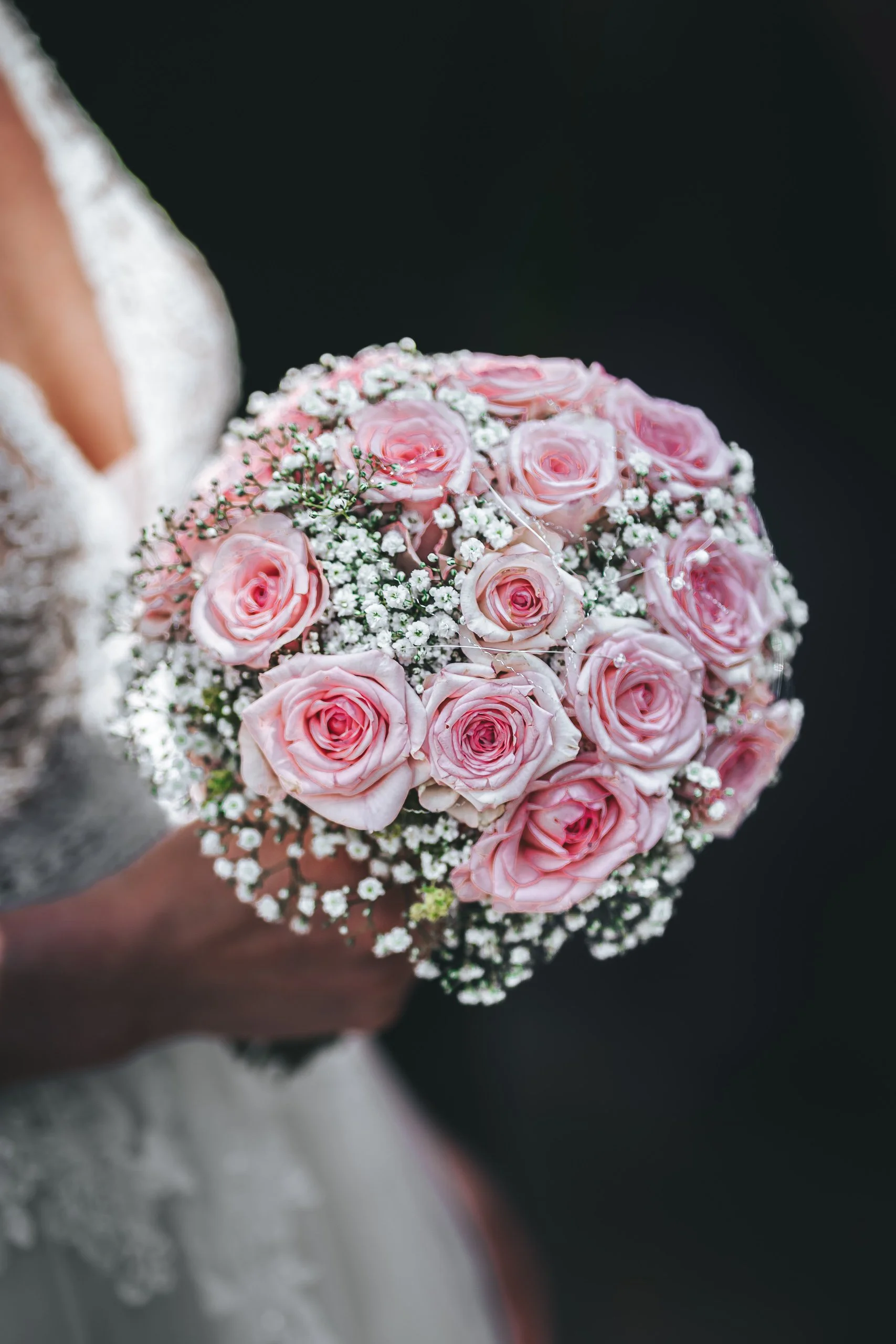 Eine Hand hält einen Brautstrauß aus rosa Rosen und kleinen weißen Blumen, vor einem dunklen Hintergrund.