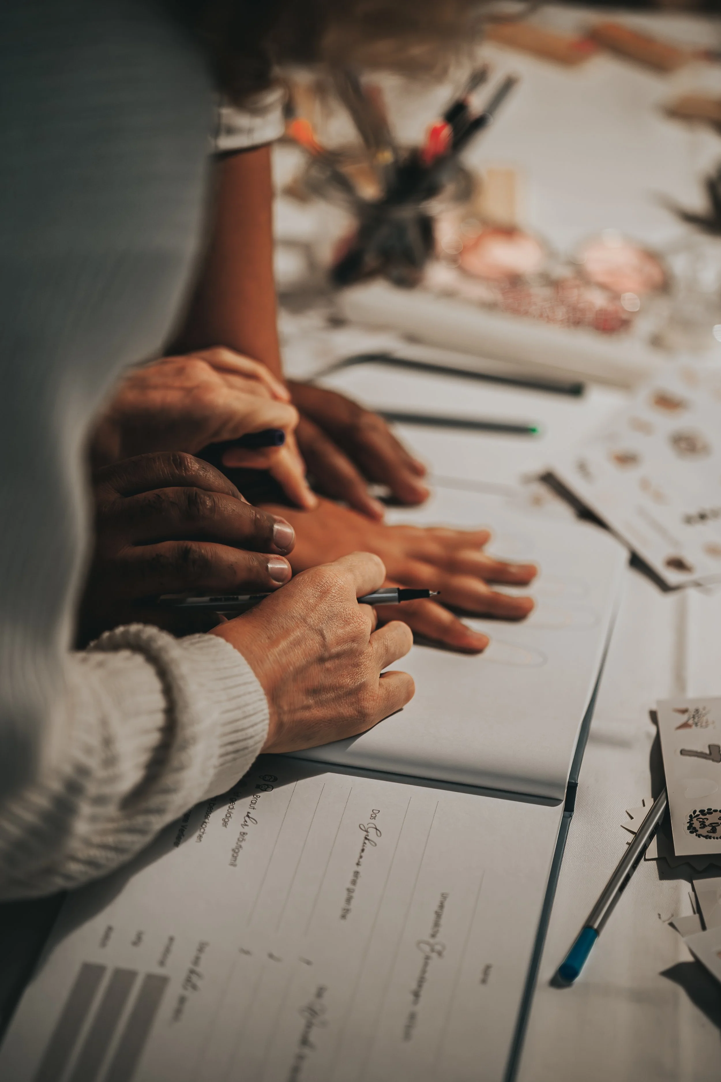 Mehrere Menschen arbeiten gemeinsam an einem Projekt, schreiben oder zeichnen auf Papier, umgeben von Stiften, Zetteln und Dekorationsmaterialien auf einem Tisch.