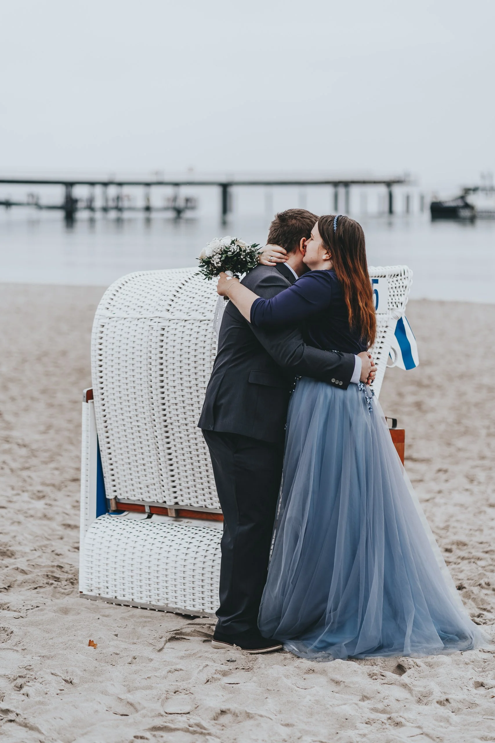 Ein Paar beim Küssen am Strand, vermutlich bei einer Hochzeit, hinter einem weißen Strandkörben, mit Wasser und Booten im Hintergrund.