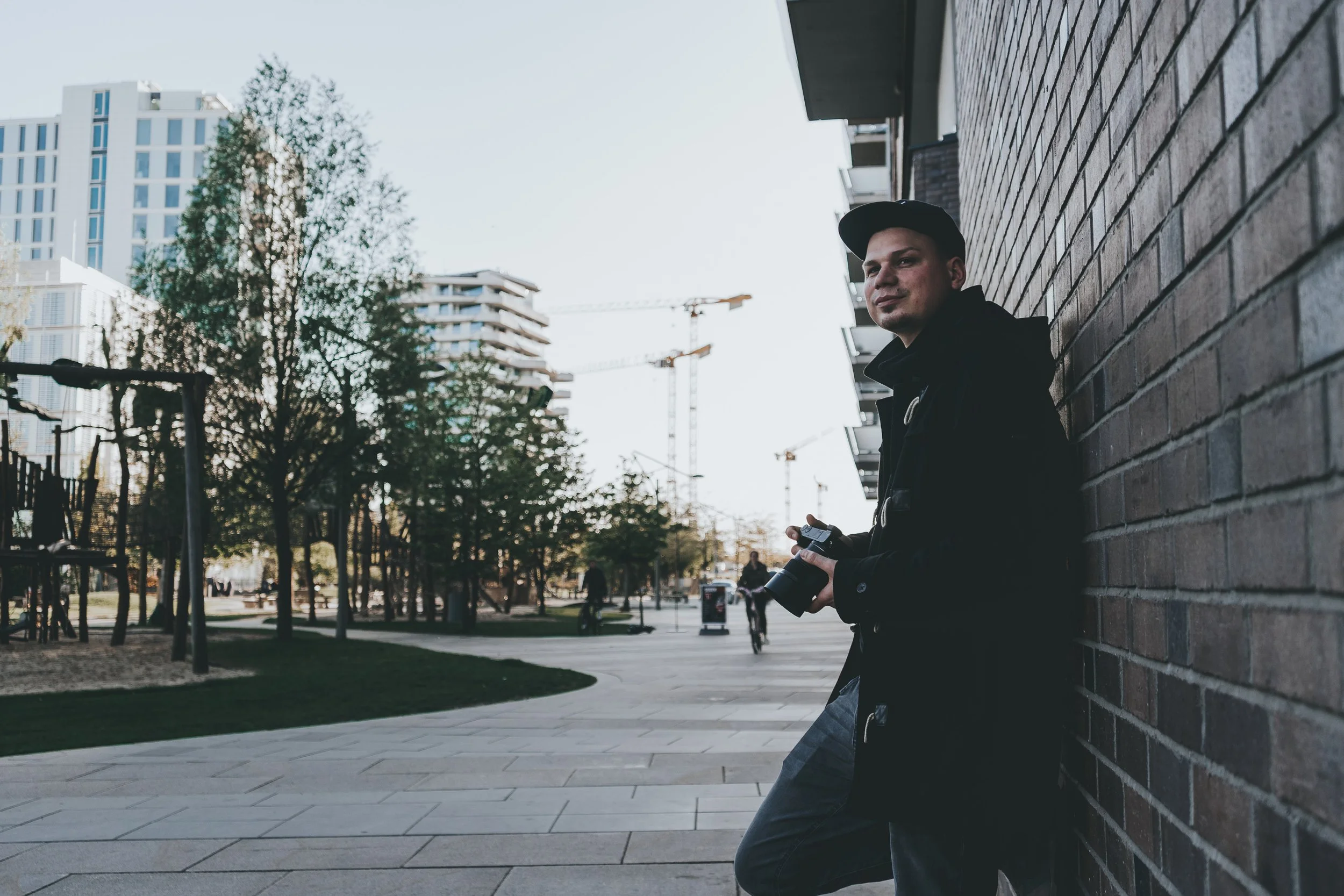 Ein junger Mann lehnt an einer Backsteinmauer im Freien, hält eine Kamera in der Hand, mit modernen Gebäuden, Bäumen und Baukränen im Hintergrund.
