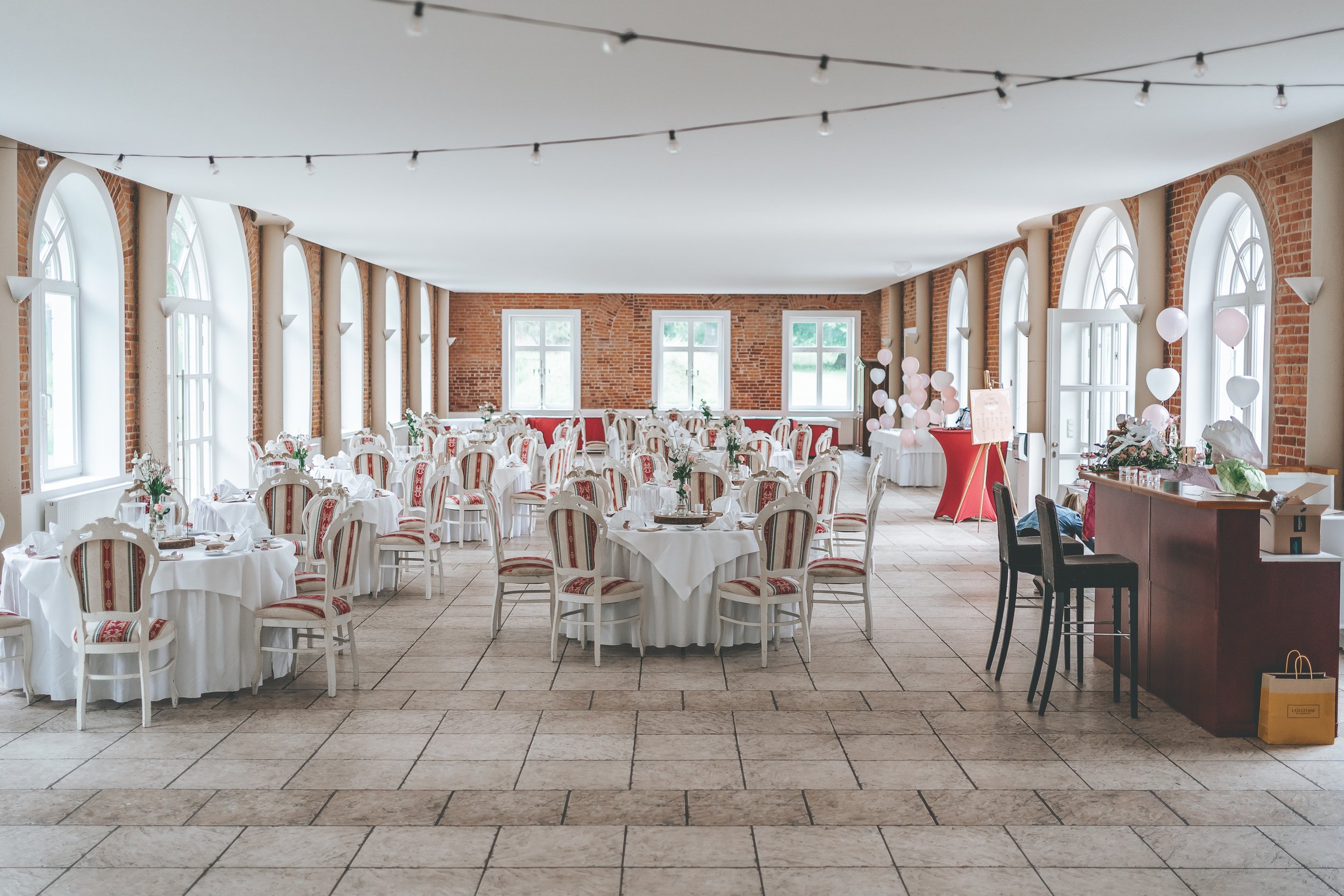 Festlich dekorierter Saal mit runden Tischen, weißem Tischtuch, Blumen und weißen Stühlen mit rotem Muster, mehrere Fenster lassen viel Tageslicht hereintreten.