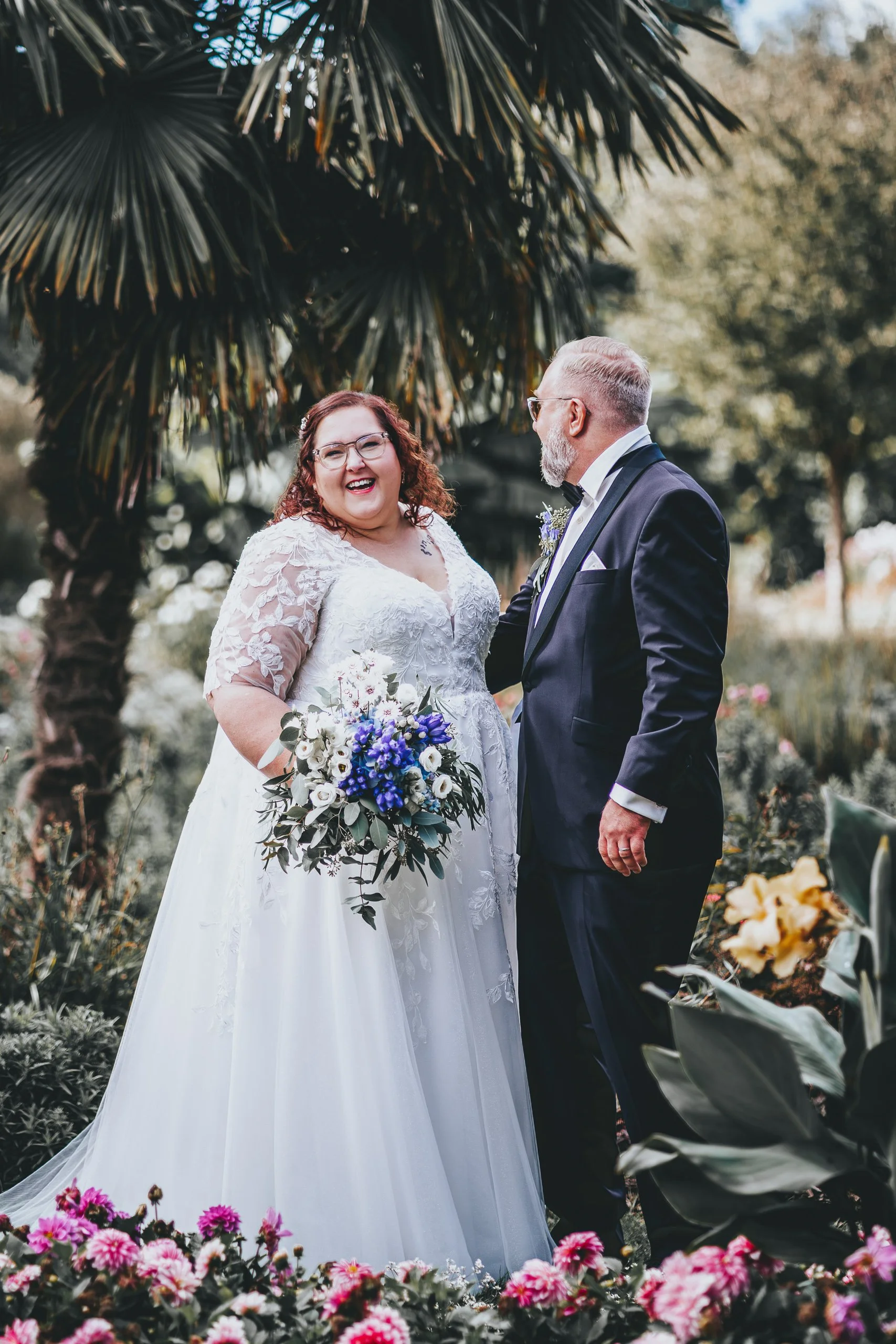 Ein lächelndes Brautpaar bei ihrer Hochzeit im Garten, die Braut trägt ein weißes Kleid mit Spitzen und hält einen Blumenstrauß, der Bräutigam trägt einen schwarzen Anzug und eine Fliege.