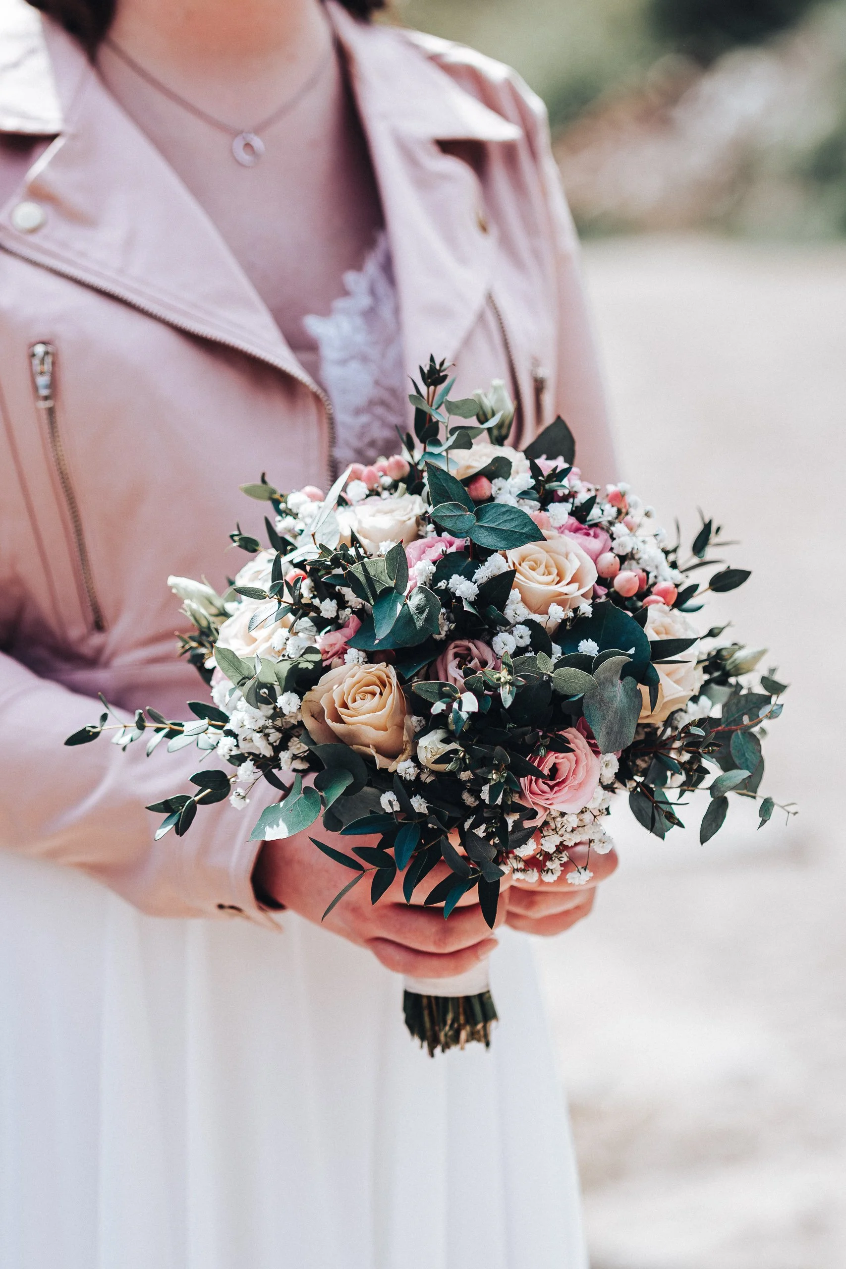 Frau in rosa Lederjacke hält einen Blumenstrauß mit weißen, rosa und cremefarbenen Blumen und grünen Blättern.