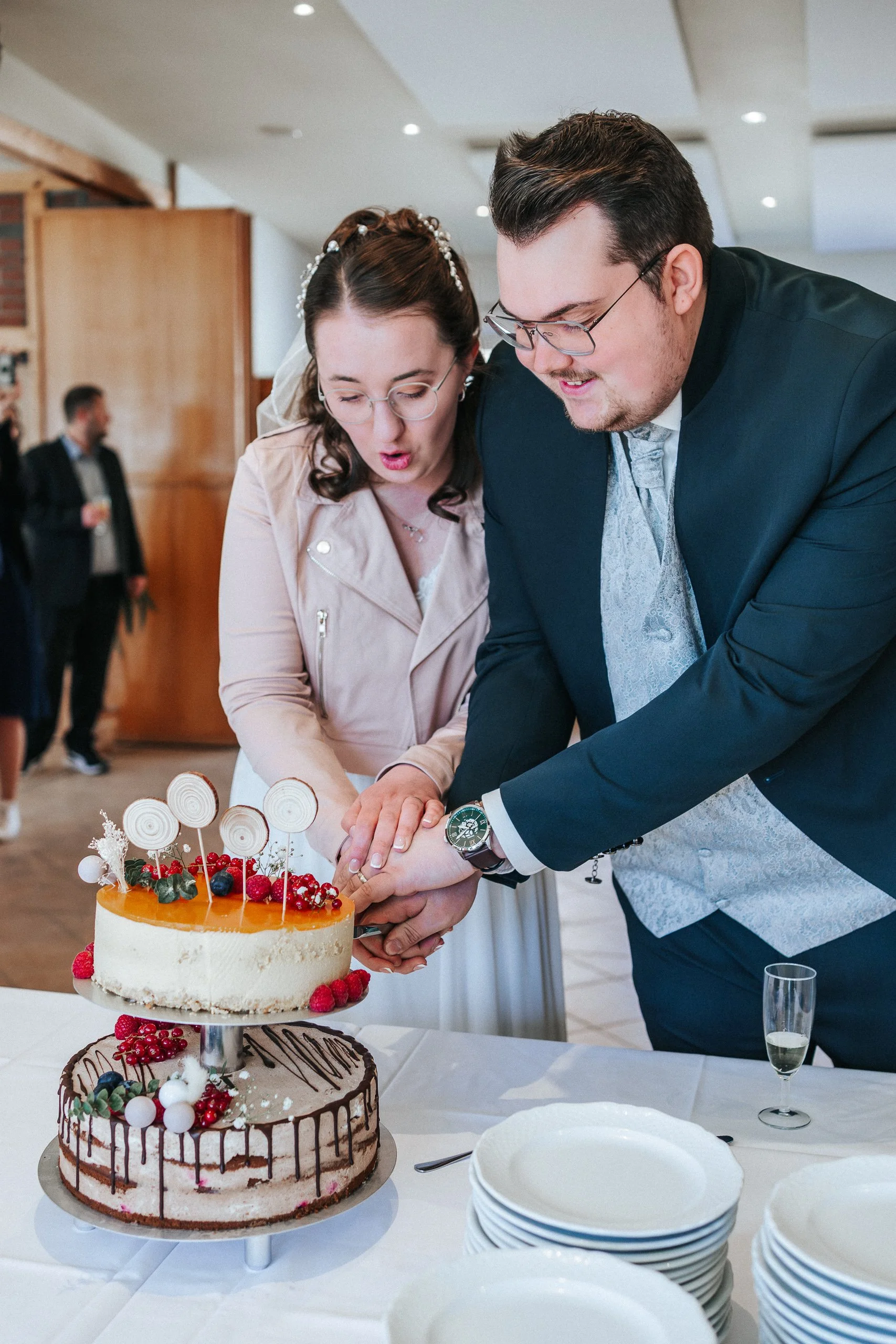 Ein Brautpaar schneidet gemeinsam eine Hochzeitstorte an. Im Vordergrund sind zwei elegante Hochzeitstorten mit Früchten und Dekorationen platziert. Die Szene findet in einem festlich dekorierten Raum statt, während im Hintergrund Gäste an der Feier 