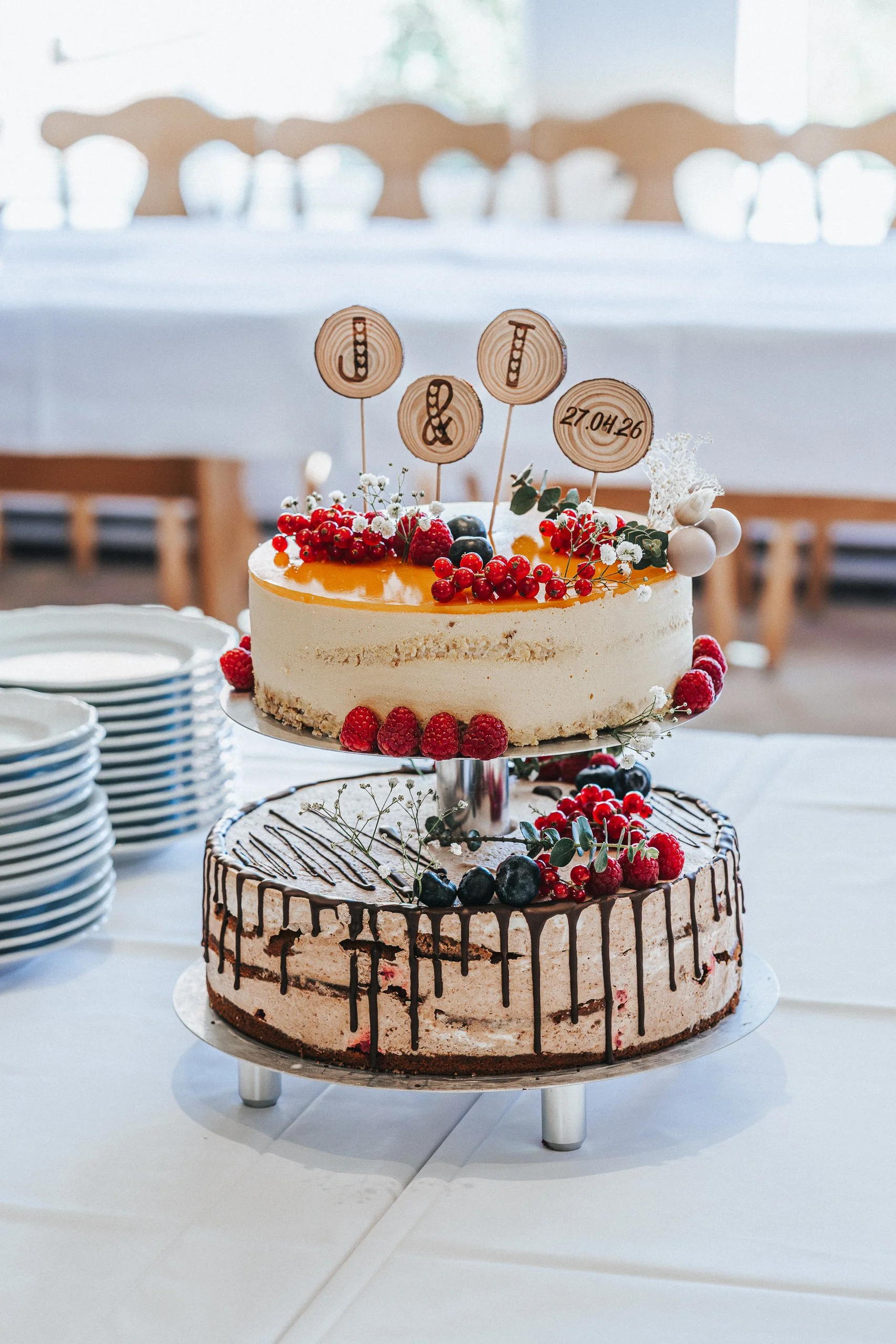 Zwei Torten übereinander, die obere mit Beeren und kleinen weißen Blumen, die untere mit Schokoladendekoration und Beeren. Auf der oberen Torte befinden sich Holzscheiben mit eingravierten Buchstaben und einer Zahl. Im Hintergrund weiße Teller. Der R