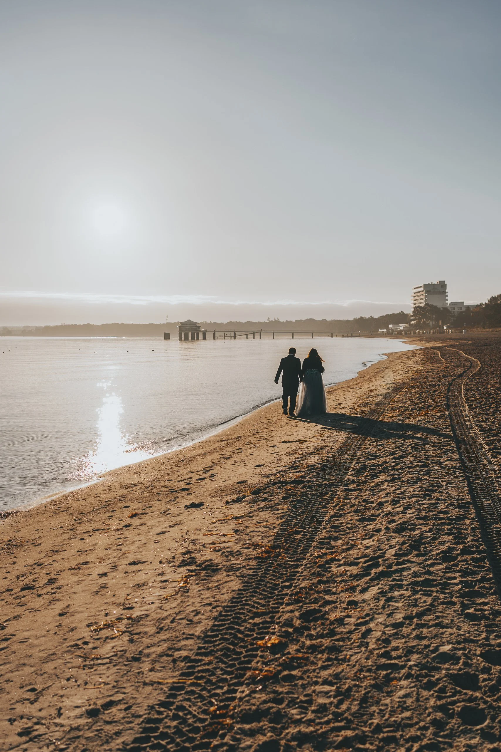 Ein Paar spaziert am Strand bei Sonnenuntergang oder Sonnenaufgang, mit einem langen Schatten, das Meer im Hintergrund und eine kleine Brücke oder Pier in der Ferne.