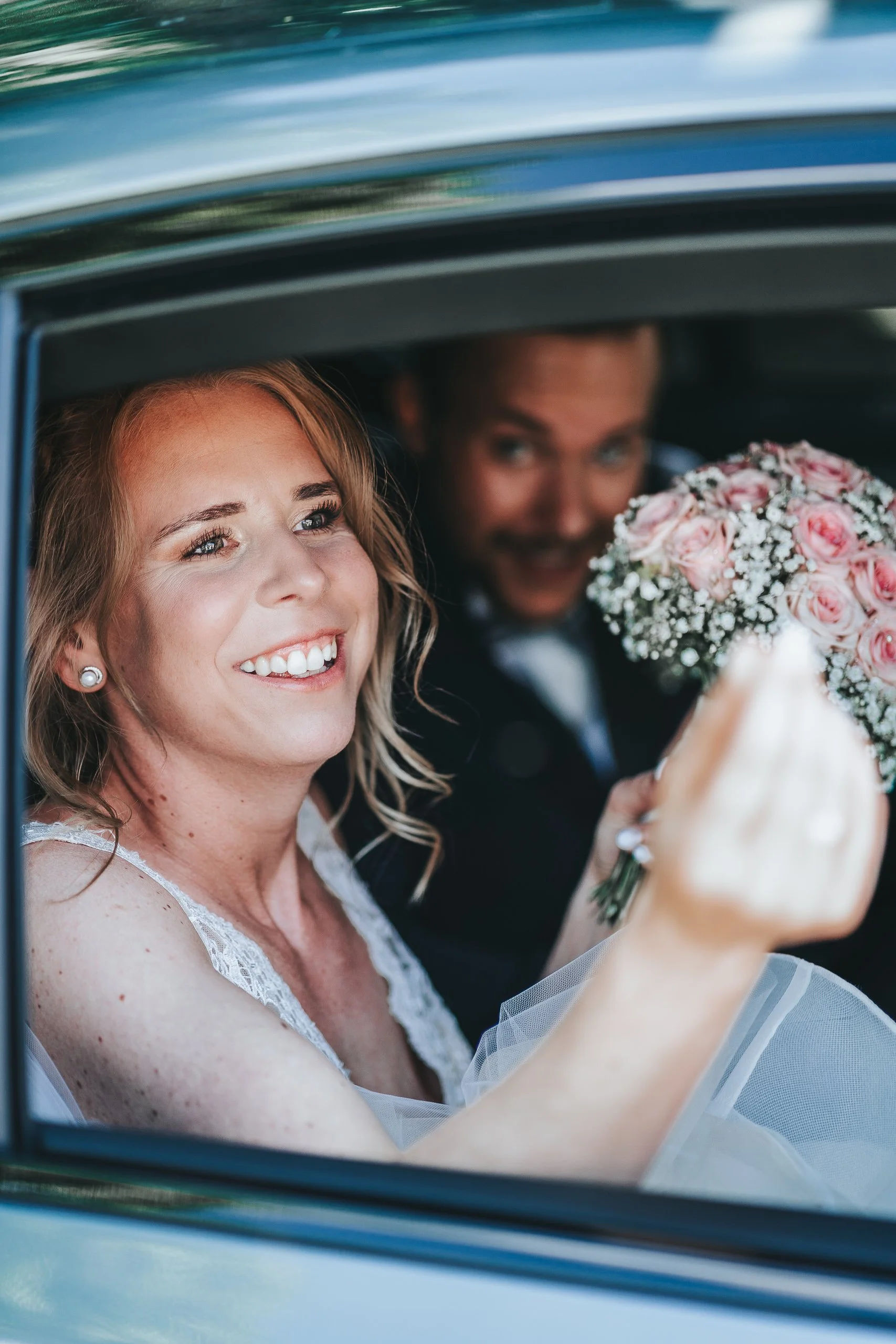 Braut und Bräutigam sitzen im Auto, die Braut lächelt und zeigt auf die Kamera, die Braut hält einen Blumenstrauß aus pinken Rosen und weißen Blumen.