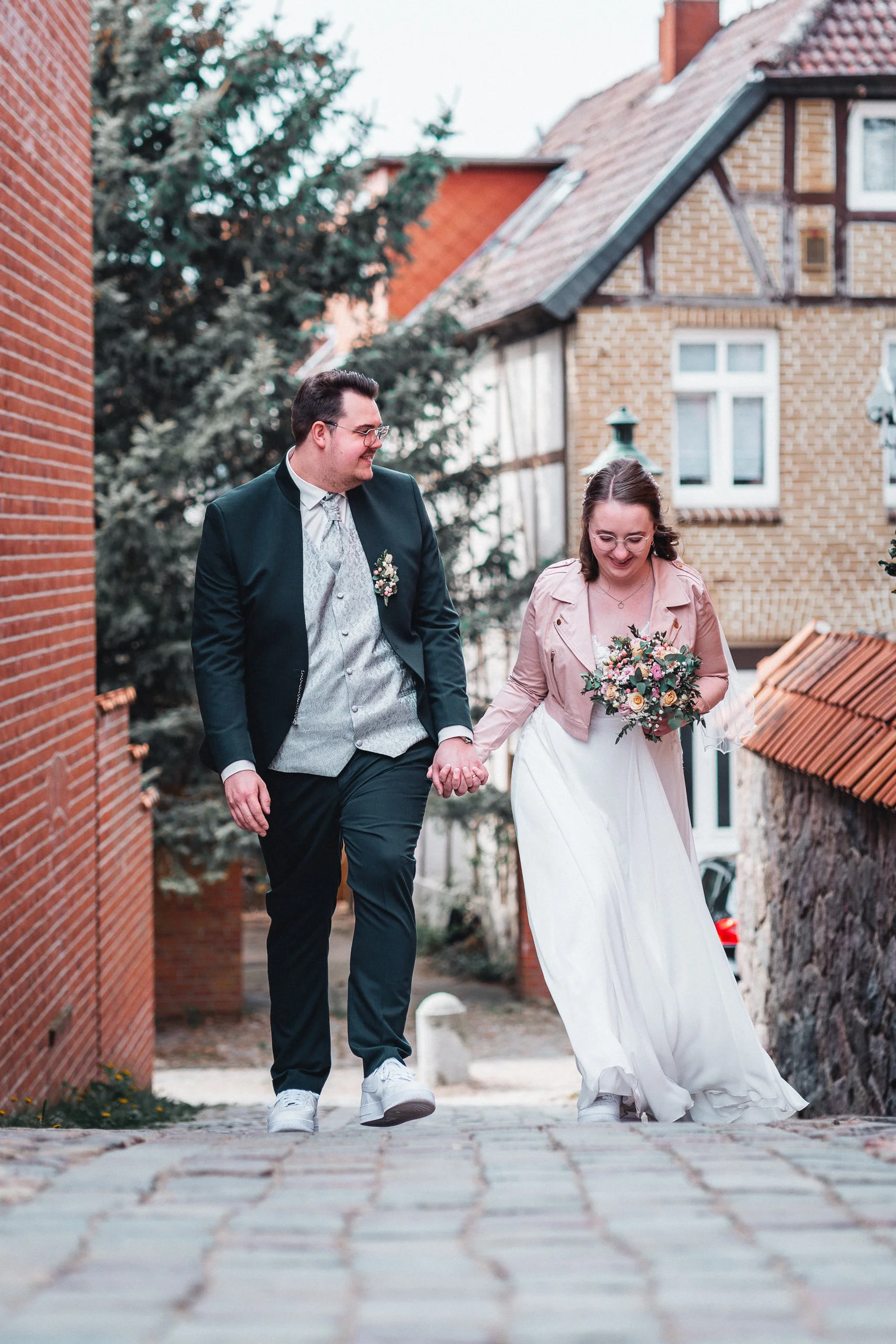Ein Brautpaar geht Hand in Hand durch eine gepflasterte Gasse in einer Altstadt. Die Braut trägt ein langes weißes Kleid mit einem Brautstrauß, der Mann trägt einen dunklen Anzug und eine Krawatte.