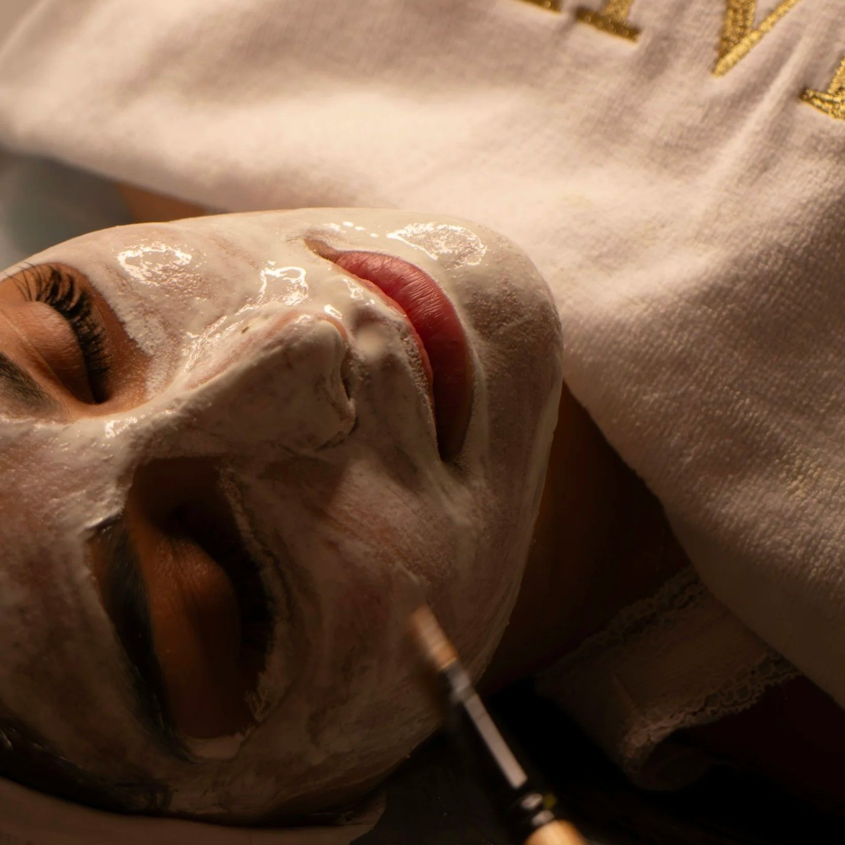 Nahaufnahme eines liegenden Gesichts mit einer Gesichtsmaskenbehandlung, die Person hat geschlossene Augen, das Gesicht ist mit cremiger Maske bedeckt, während eine Hand einen Pinsel benutzt, um die Maske auf das Gesicht aufzutragen.