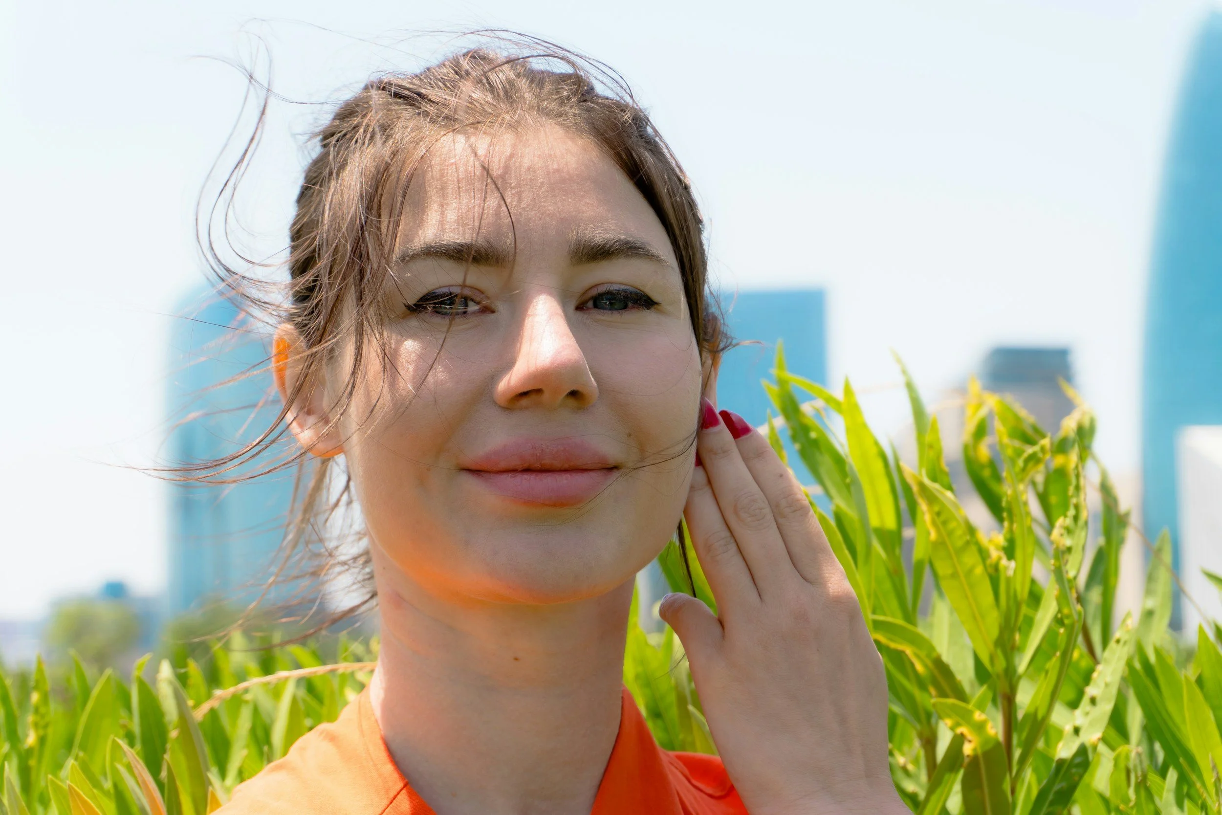 Junge Frau mit braunen Haaren und rotem Lippenstift, posaunt im Feld, hinter ihr zahlreiche grüne Pflanzen, hohe Gebäude im Hintergrund, Himmel ist klar und blau.