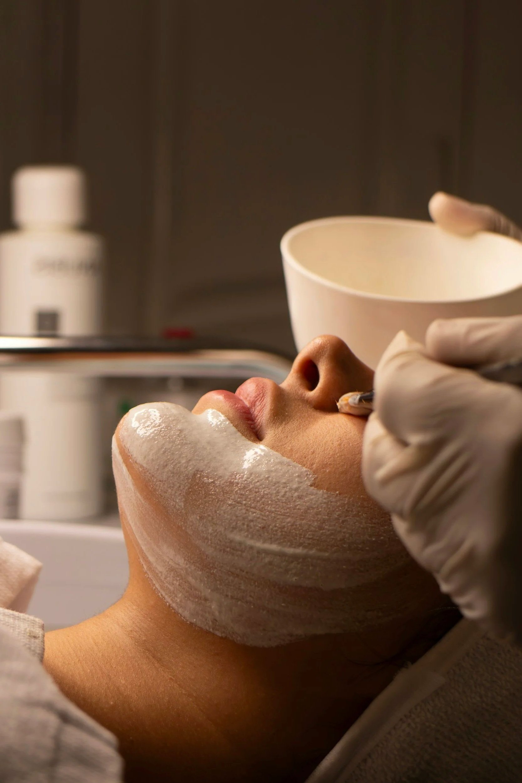 Eine Person erhält eine Gesichtshautpflegebehandlung im Spa, mit einer weißen Maske im Gesicht und einer Schüssel, die wahrscheinlich mit Pflegeprodukten gefüllt ist, in der Hand einer Person mit Handschuhen.