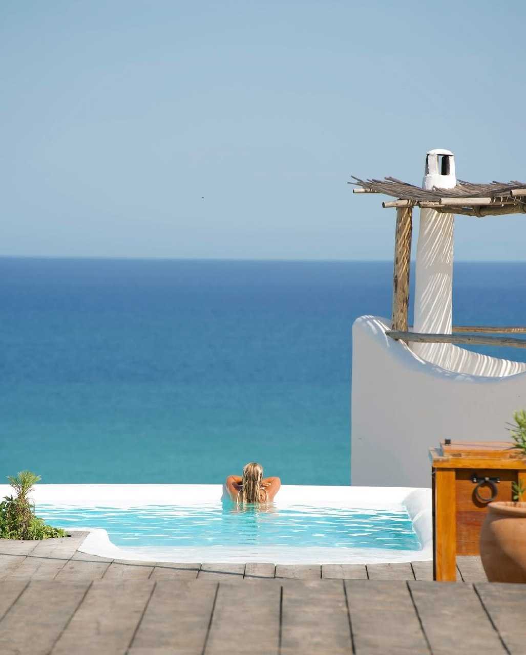 Person relaxing in infinity pool overlooking ocean, with rustic wooden deck and a simple canopy structure nearby.