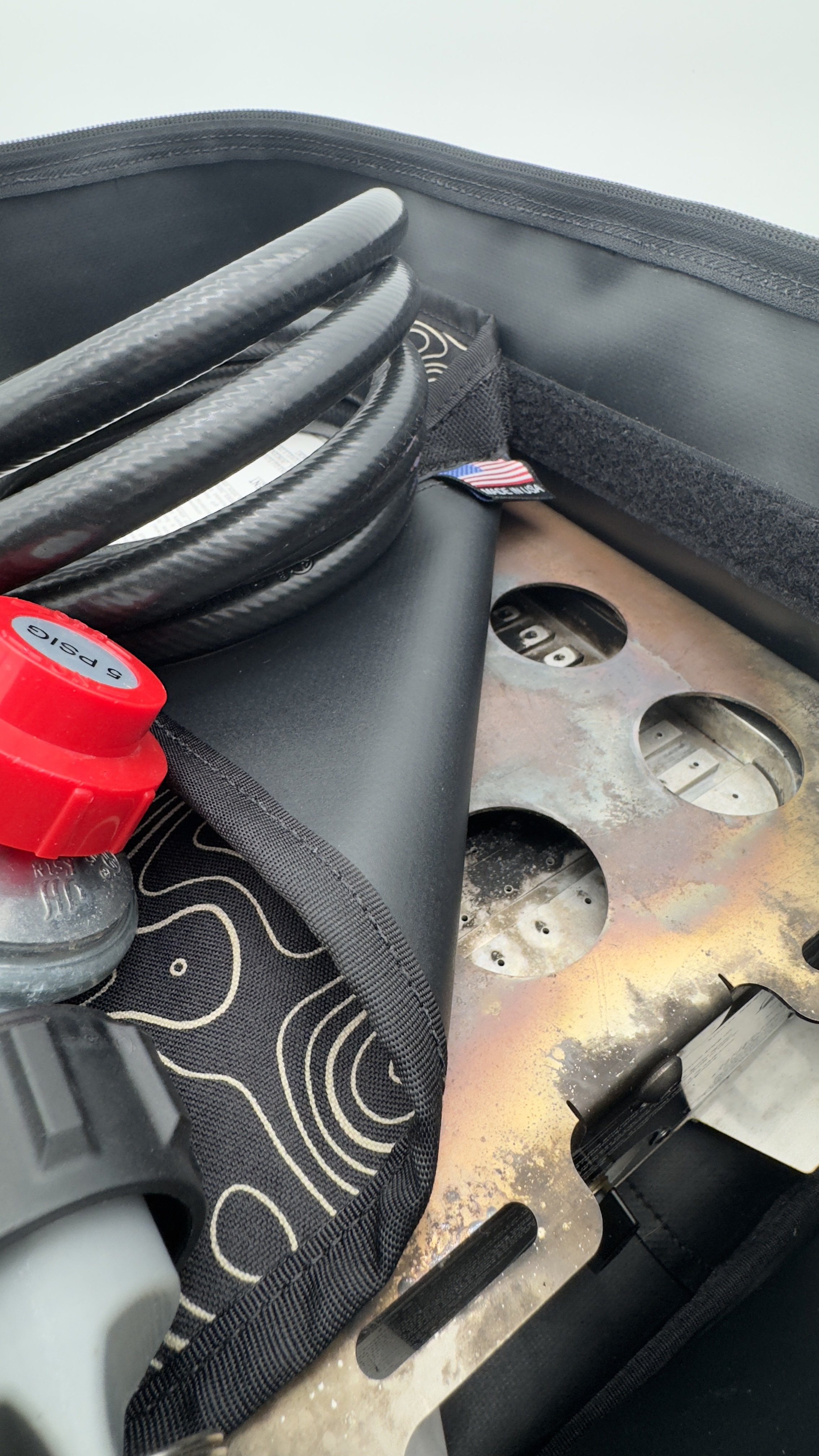 Close-up of a storage bag with coiled propane hose, a red propane regulator, and a propane fire pit with circular cutouts.