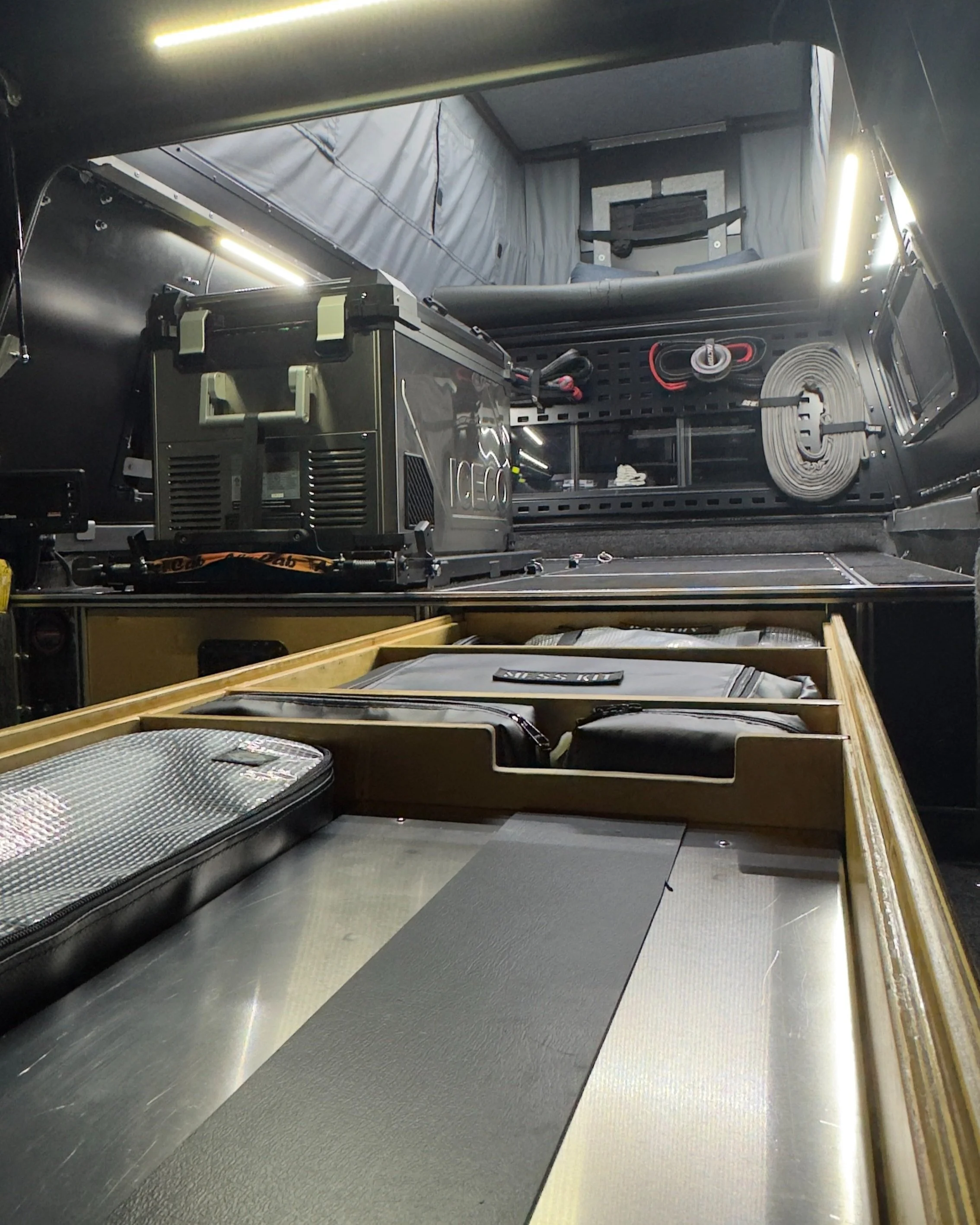 Interior view of a camper or van with various equipment, including a gray fridge freezer, black boxes, and organized supplies, with overhead lighting and storage compartments.