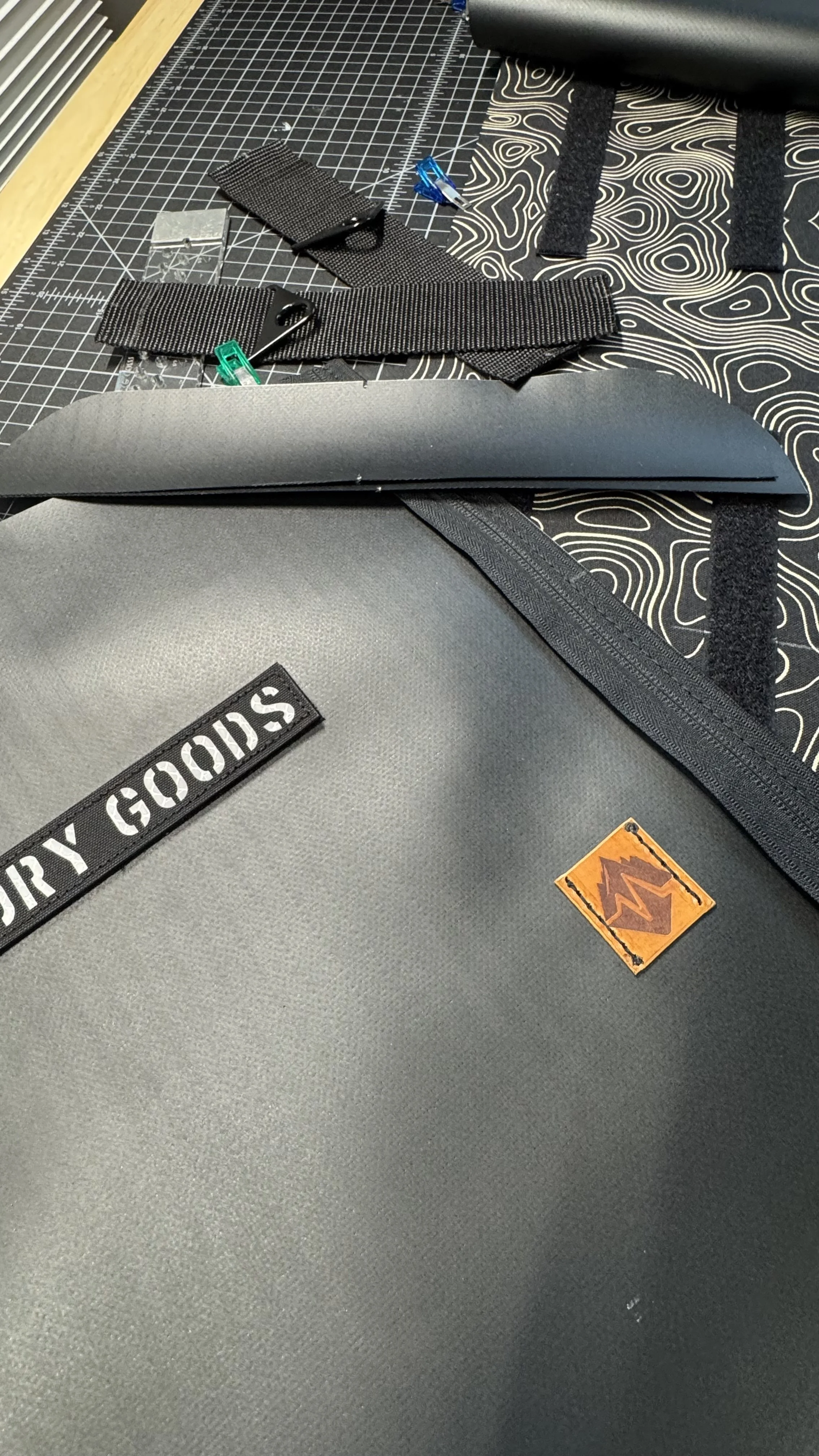 Close-up of a grey dry bag with black zipper, branding label, and orange logo, placed on a black patterned surface with various gear items including straps, clips, and a cutting mat with measurement grid.