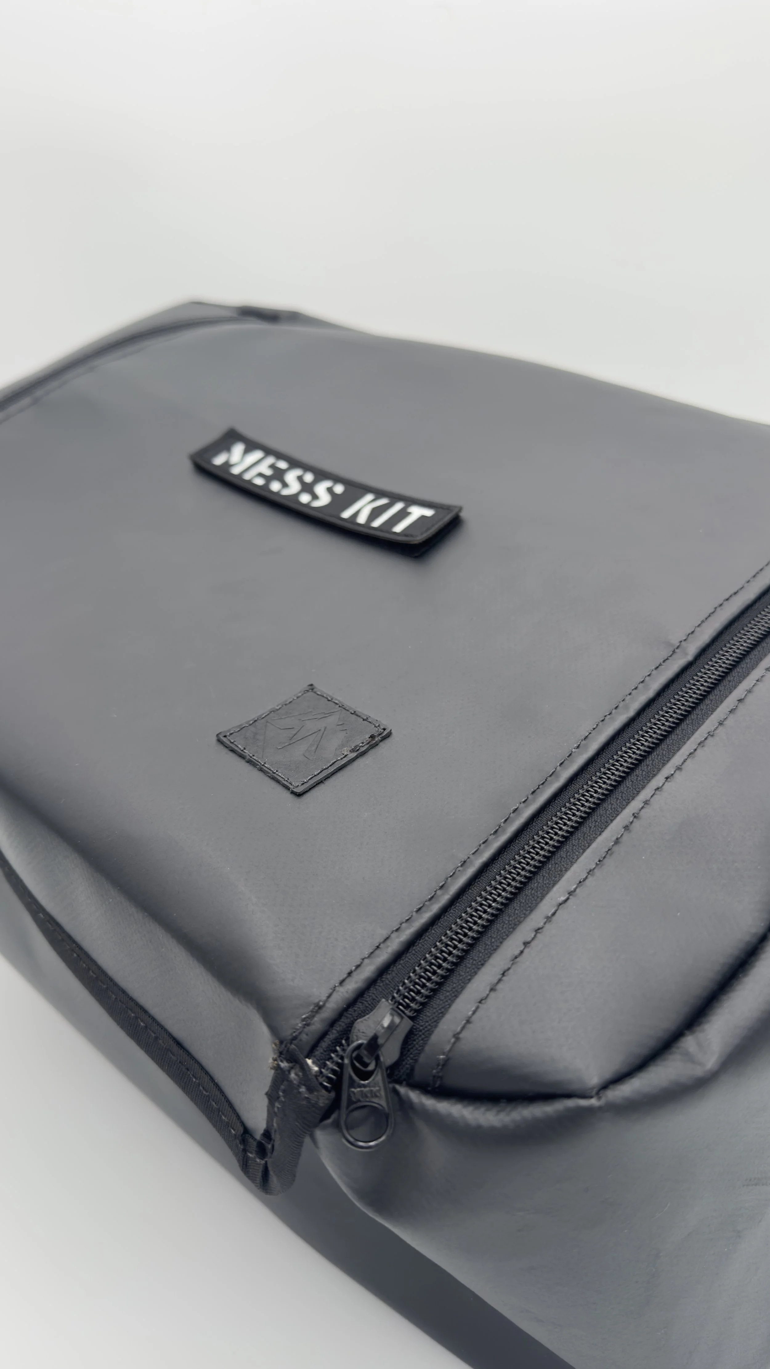 A black bag with a zipper, a label that reads "MESS KIT," and a small embossed logo on the front.