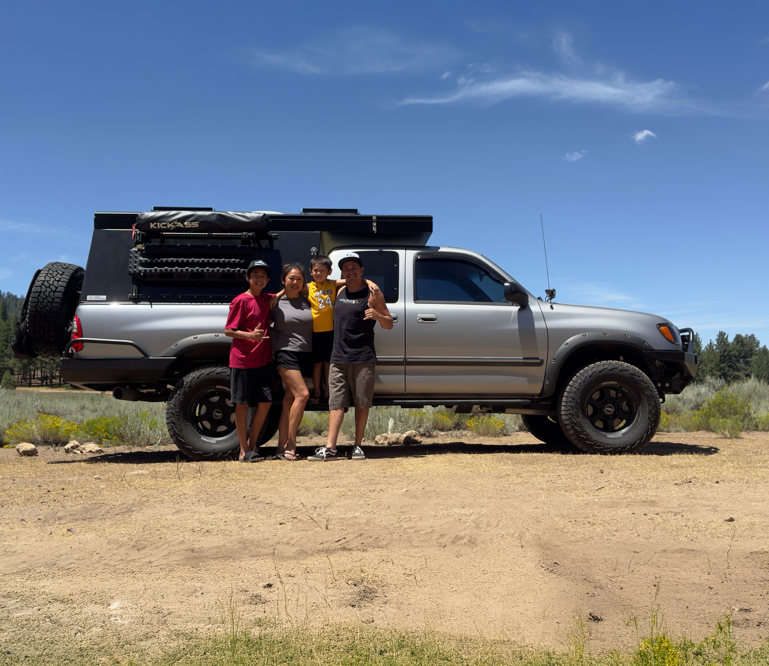 Four people standing beside a silver off-road vehicle on a dirt trail in a rural area with trees in the background. Clear sky above.