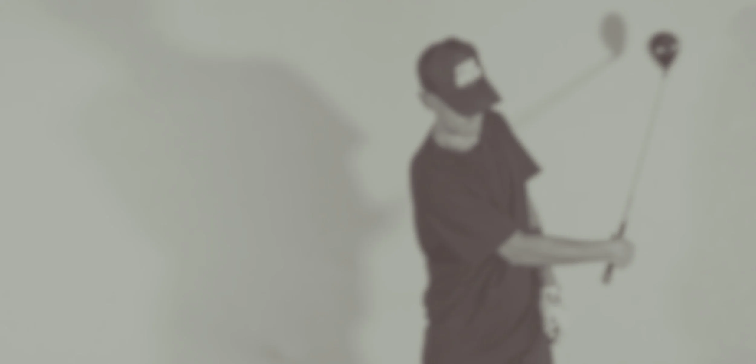 A man with a beard wearing a dark t-shirt and glasses, holding a selfie stick with two balloons attached, standing against a plain white wall.