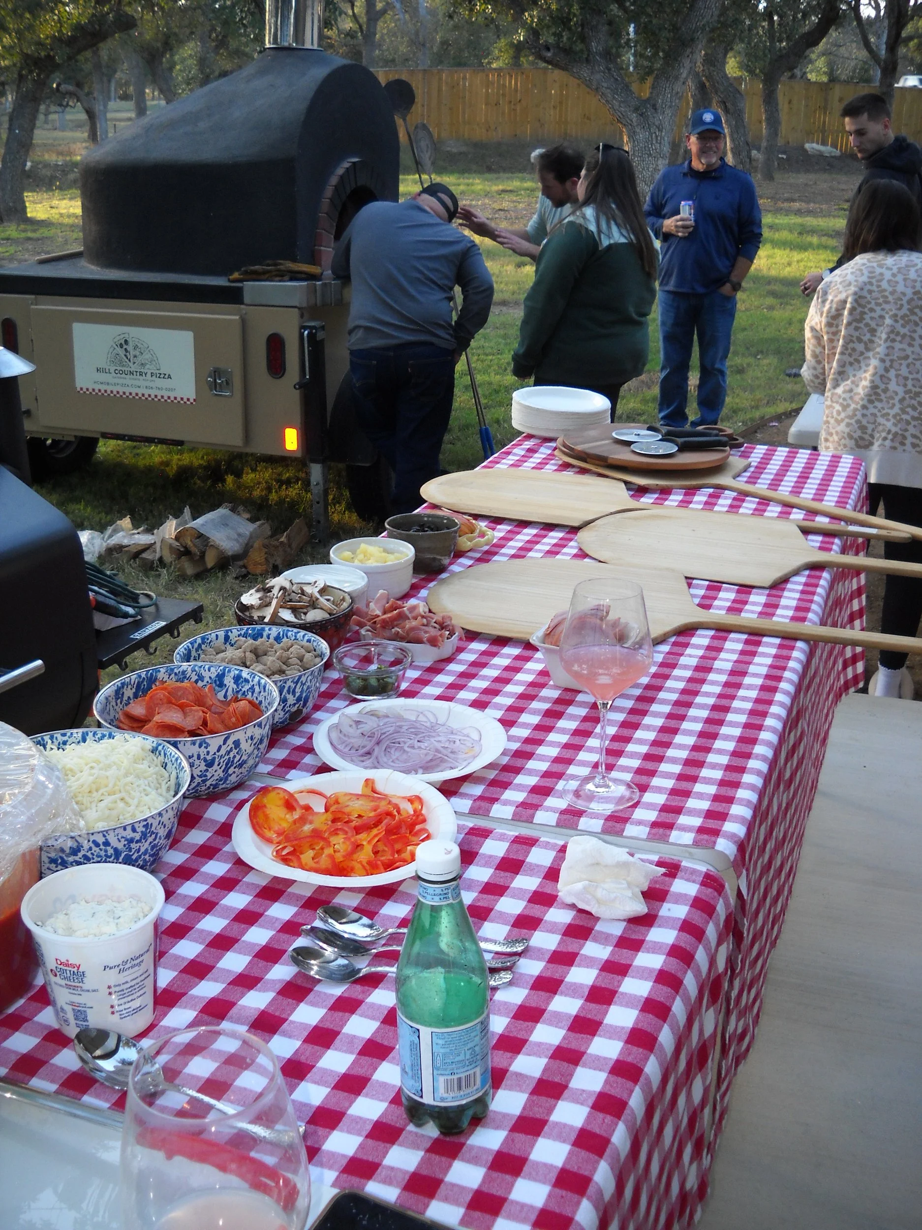 An outdoor barbecue with a table covered in a red and white checkered tablecloth displaying various ingredients like sliced tomatoes, onions, shredded cheese, and meats, with a pizza oven in the background and people gathered around, some preparing food and others talking, with trees and a wooden fence in the background.