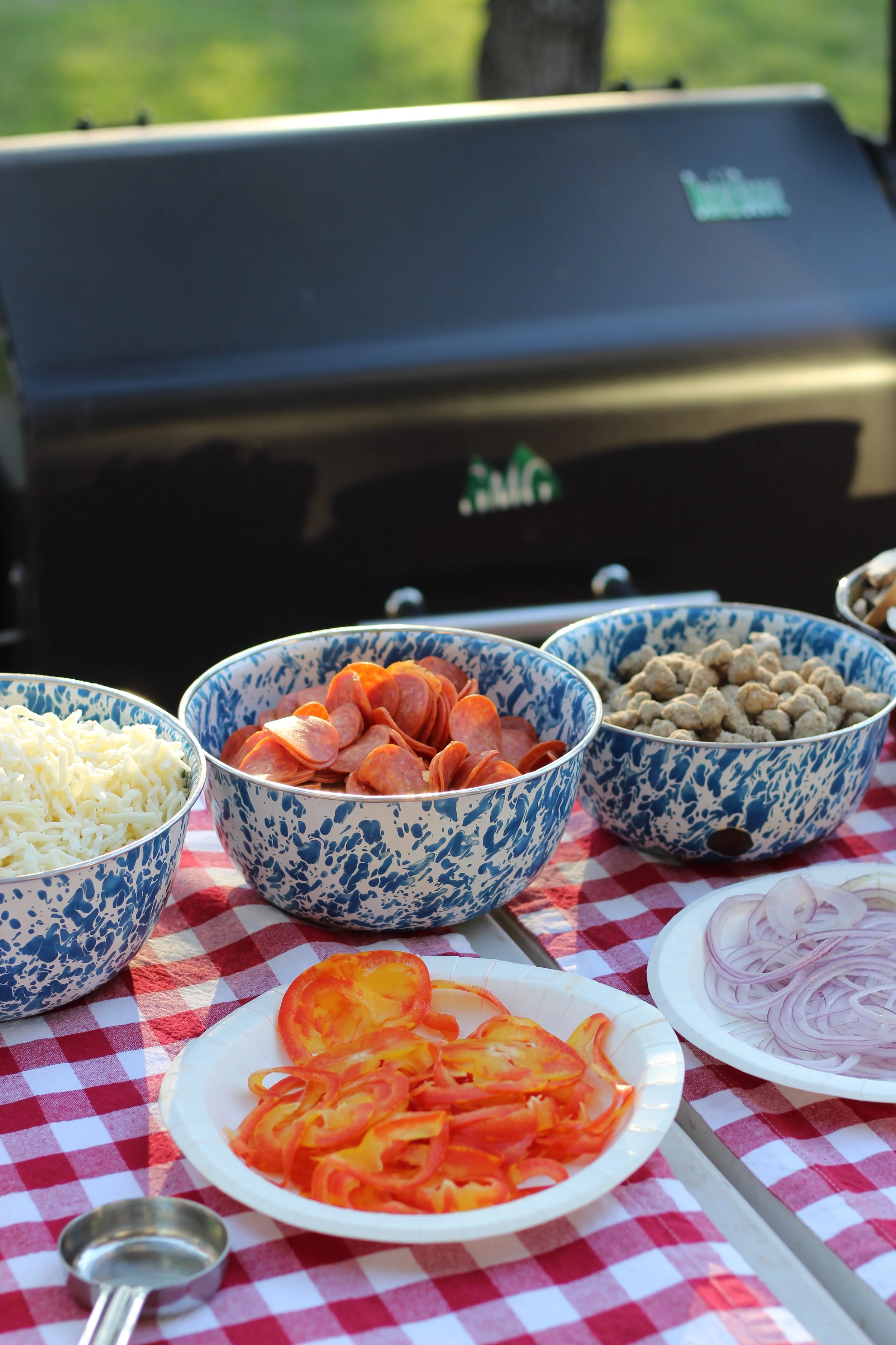 Bowls of sliced tomatoes, onions, shredded cheese, pepperoni, and crumbled sausage on a red and white checkered tablecloth in front of a black grill.