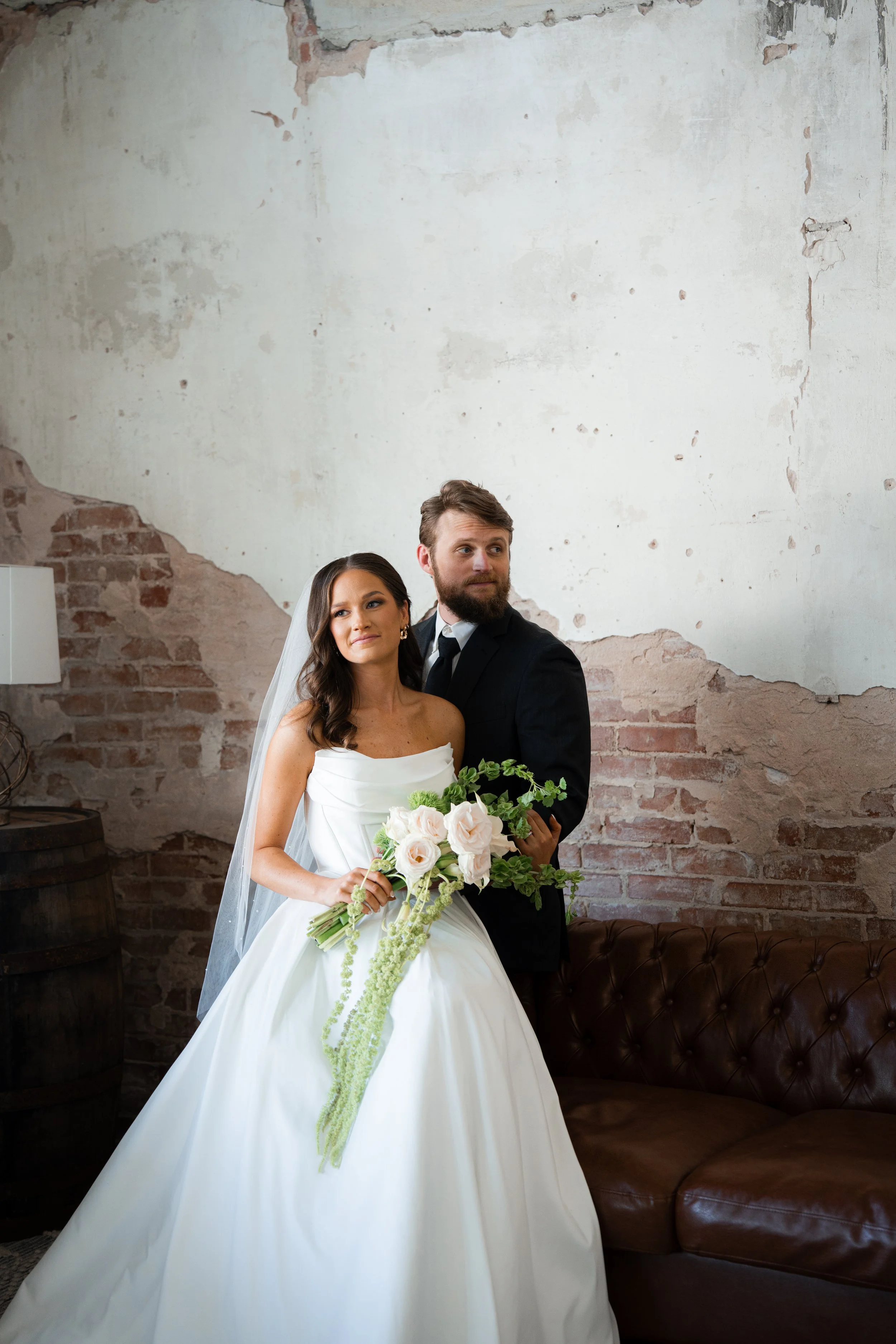 Bride and groom pose indoors with a rustic brick wall and leather couch in the background. The bride wears a strapless white wedding gown and holds a bouquet of white roses and greenery. The groom wears a black suit and white shirt.