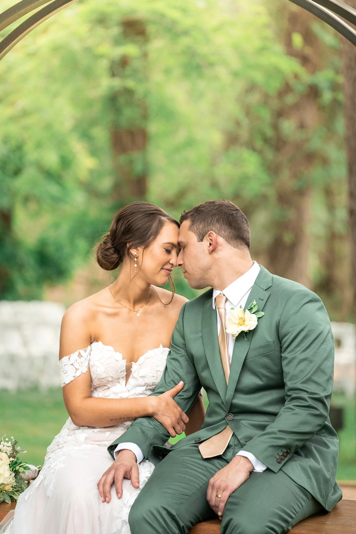 A bride and groom with foreheads touching, sitting outdoors in a wooded area on their wedding day, dressed in wedding attire.