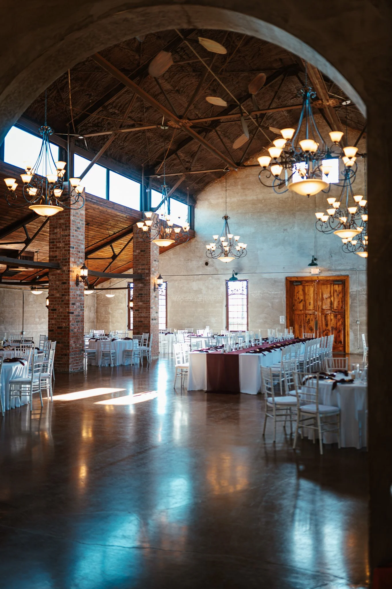 Elegant banquet hall with high wooden ceilings, chandeliers, brick pillars, and set dining tables for an event.