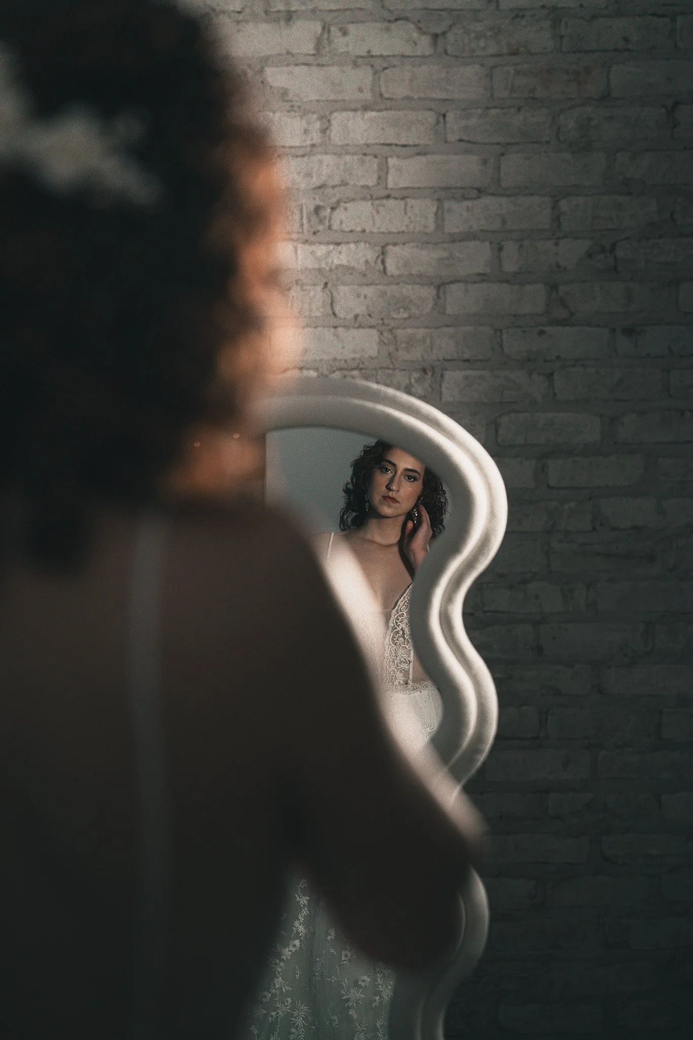 A woman in a white dress looking at her reflection in a uniquely shaped mirror, with a brick wall in the background.