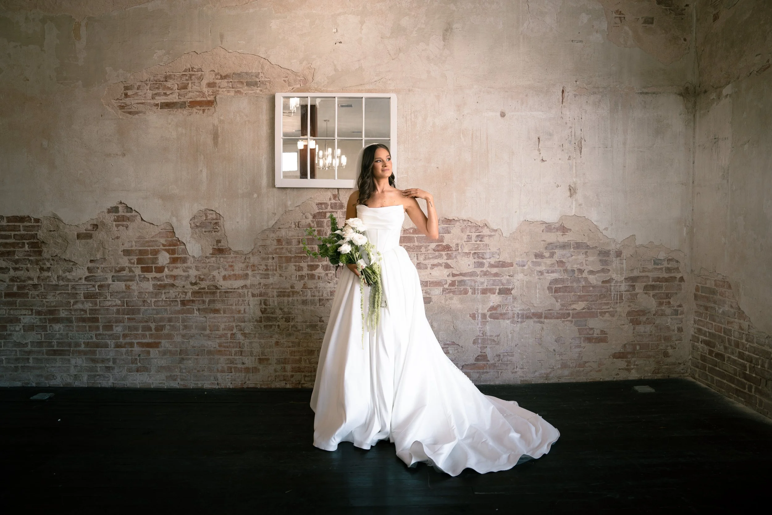 A bride in a white wedding gown holding a bouquet of white flowers and greenery, standing indoors against a weathered brick and plaster wall with a window and chandelier reflection visible in the mirror.