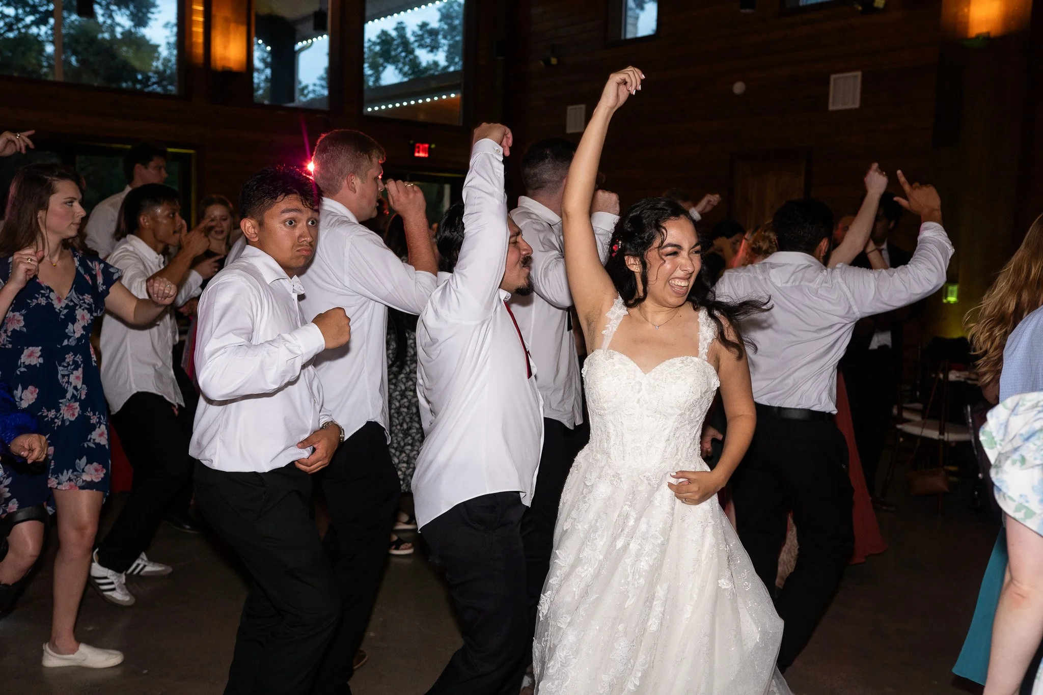 People dancing at a wedding reception, with the bride in a white dress and the guests in formal and semi-formal attire, in a wooden venue with large windows.
